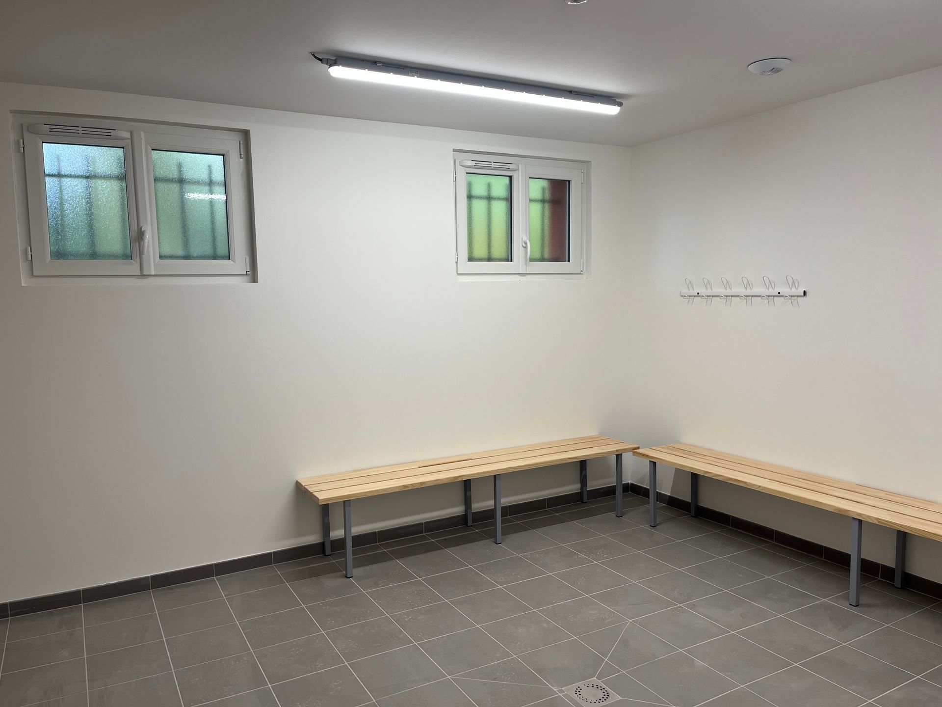 Un couloir étroit carrelé, avec un mur blanc à gauche et des fenêtres à stores orange à droite, menant à une porte.