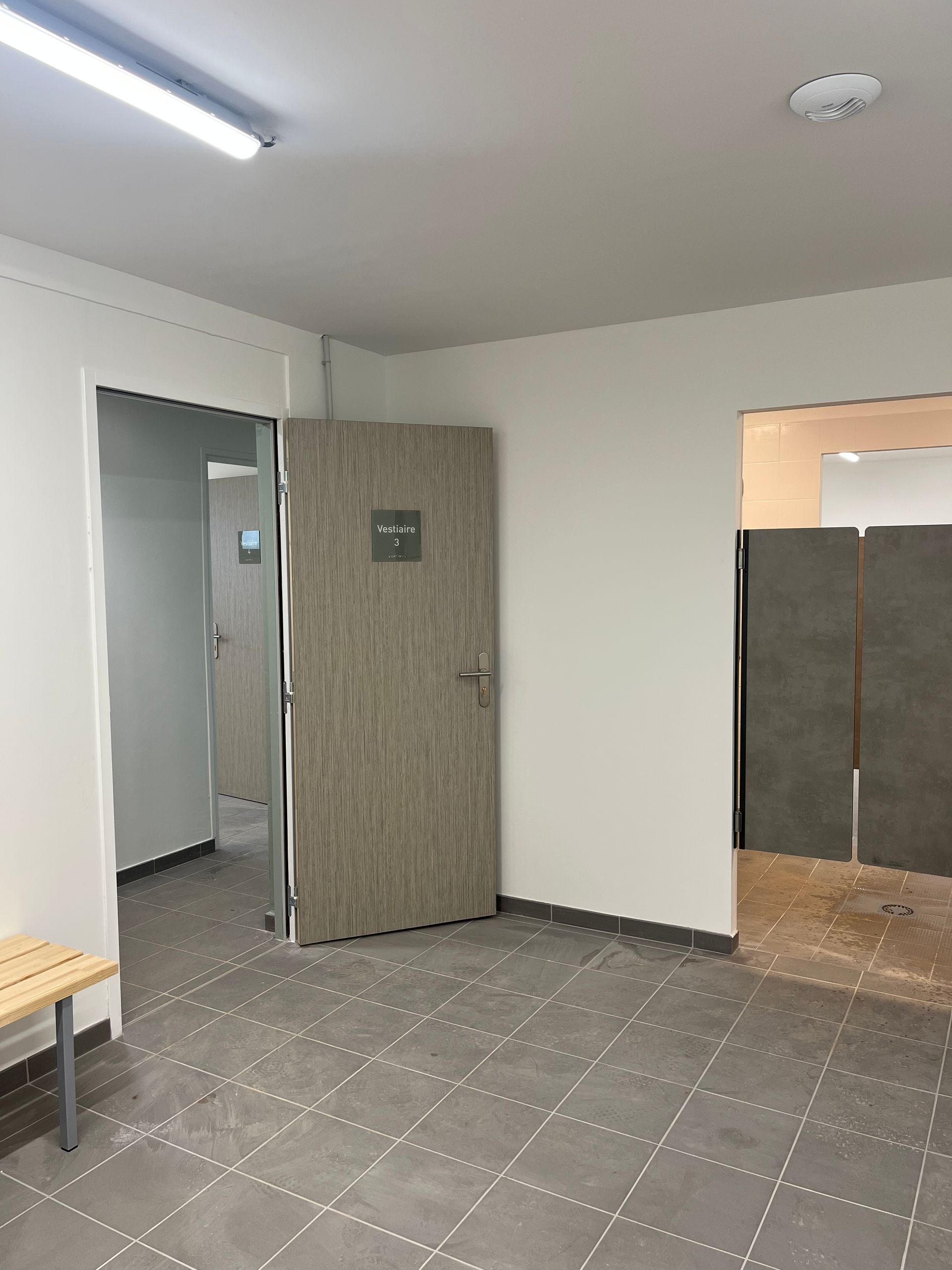 Un vestiaire avec des rangées de casiers bleus, verts et violets, de la moquette grise et deux bancs en bois au centre.