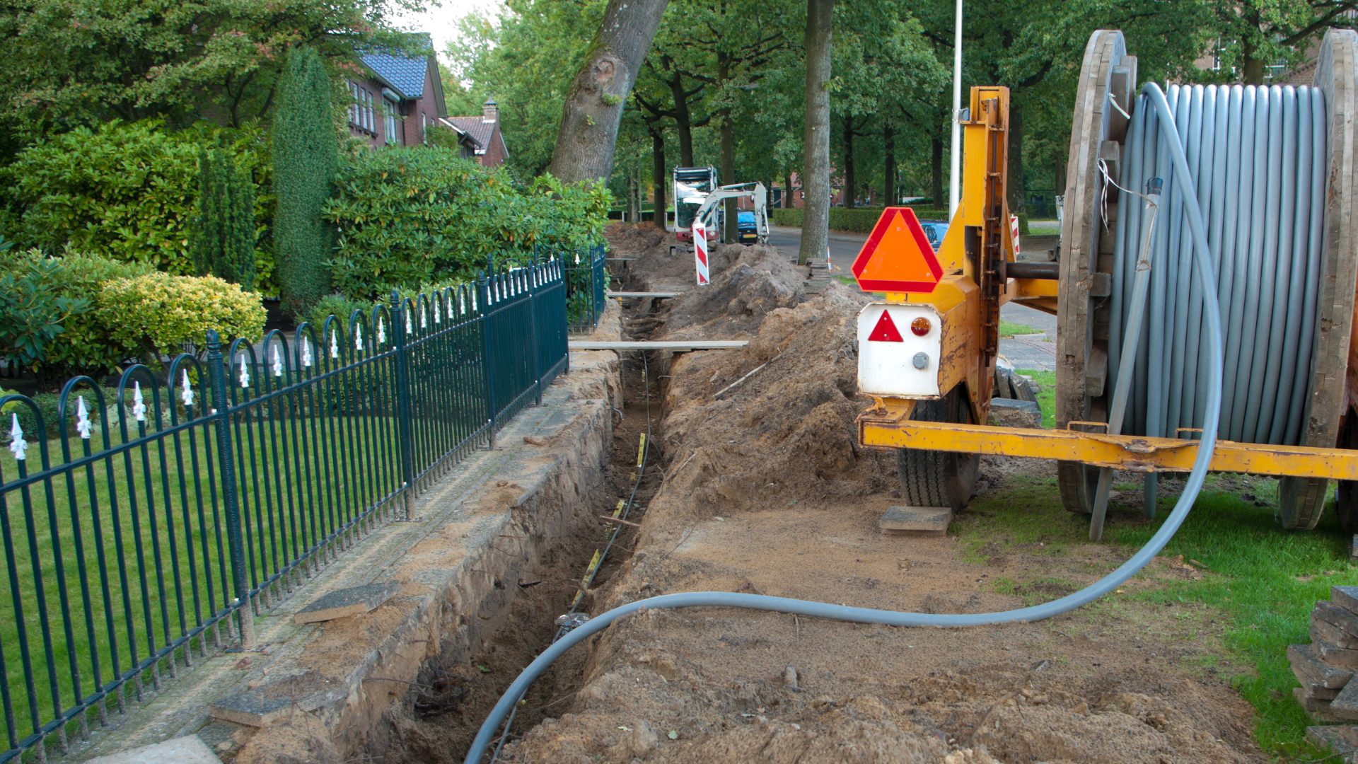 Une tranchée est en cours de creusement le long d'un trottoir, à côté d'une clôture résidentielle, avec un grand tambour de câble sur une remorque à proximité.