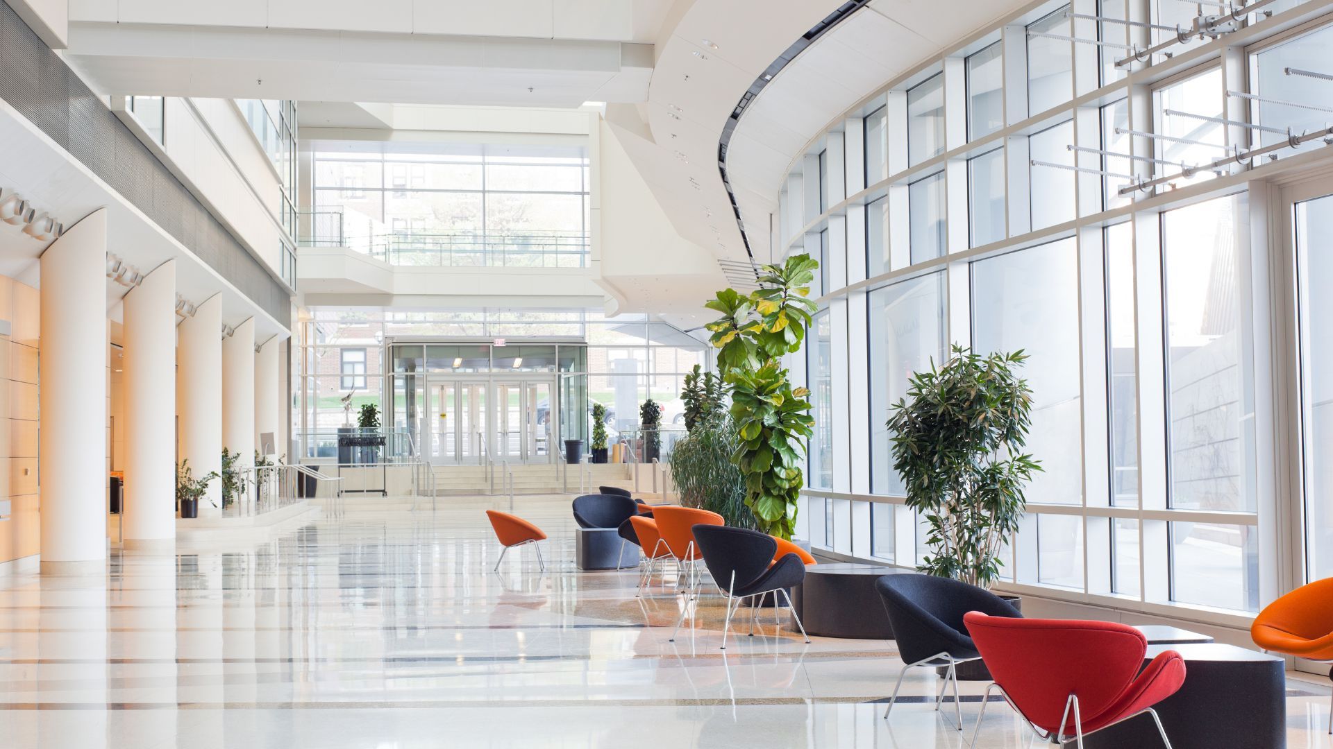 Un hall d'entrée lumineux et moderne avec des baies vitrées, des piliers blancs, des plantes en pot et des fauteuils colorés.