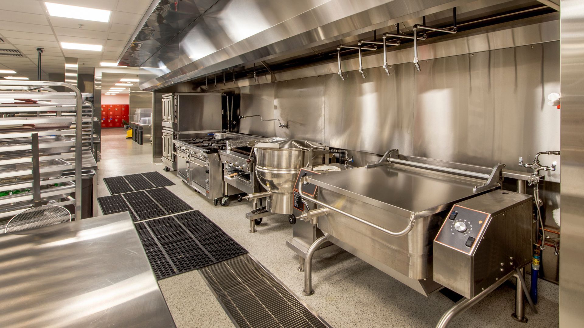 Une cuisine commerciale professionnelle équipée de matériel en acier inoxydable, d'une poêle basculante et d'une cuisinière sous une hotte aspirante.