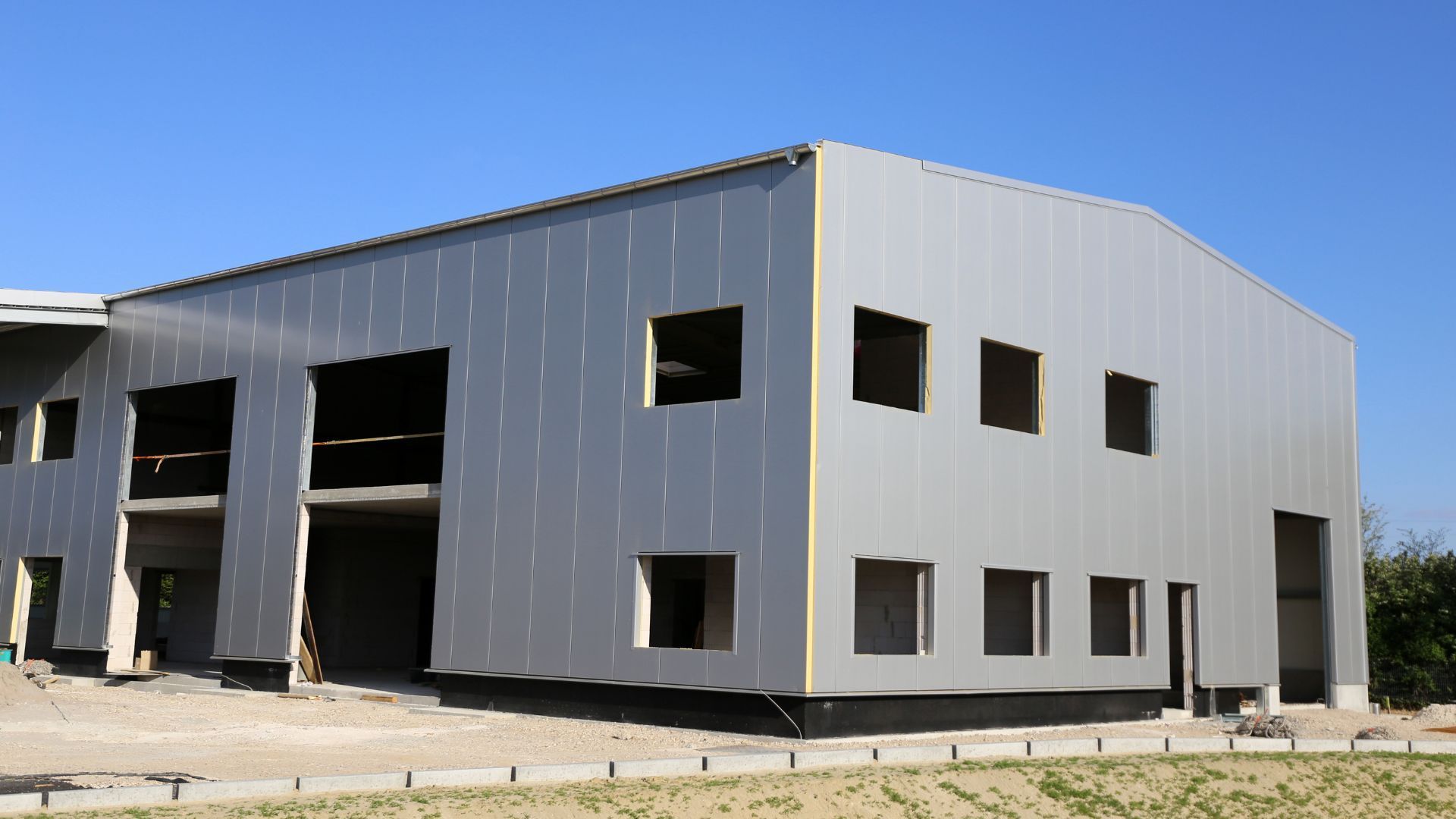 Un bâtiment industriel moderne en métal gris, inachevé, se détachant sur un ciel bleu clair, laissant apparaître des fenêtres et des travées ouvertes.