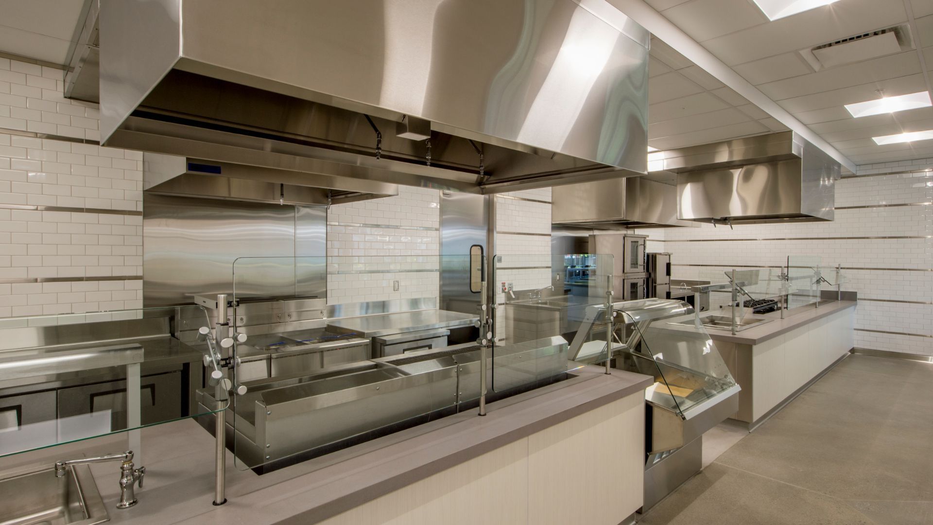 Cuisine commerciale moderne avec équipements en acier inoxydable, hottes aspirantes et murs carrelés blancs.