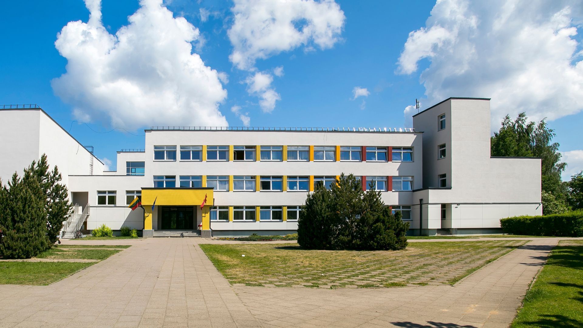 Un bâtiment scolaire de trois étages, de couleur claire, sous un ciel d'un bleu éclatant, avec une entrée jaune et une cour pavée.