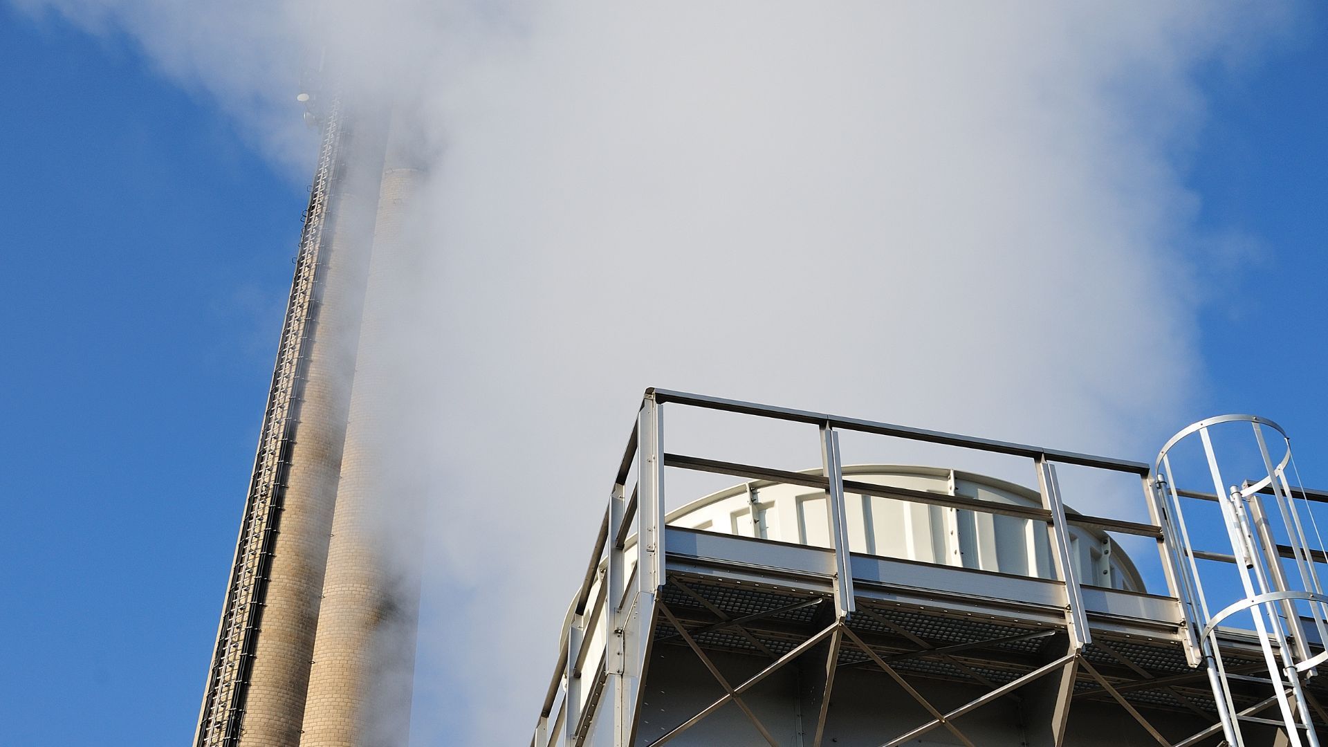 Une haute cheminée en briques crachant une épaisse vapeur blanche se dresse à côté d'une tour de refroidissement métallique, sur fond de ciel bleu clair.