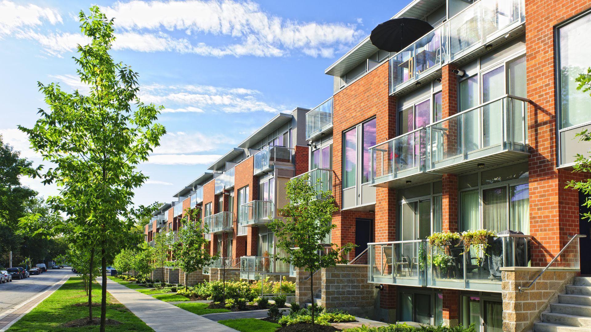 Une rangée de maisons de ville modernes en briques, avec des balcons vitrés, borde une rue avec un trottoir et des arbres verts sous un ciel bleu.