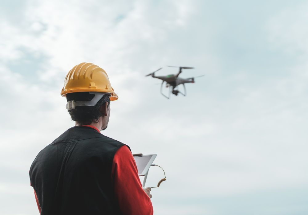 Un ouvrier du bâtiment, coiffé d'un casque jaune, pilote un drone à l'aide d'une tablette tactile, sur fond de ciel lumineux et nuageux.