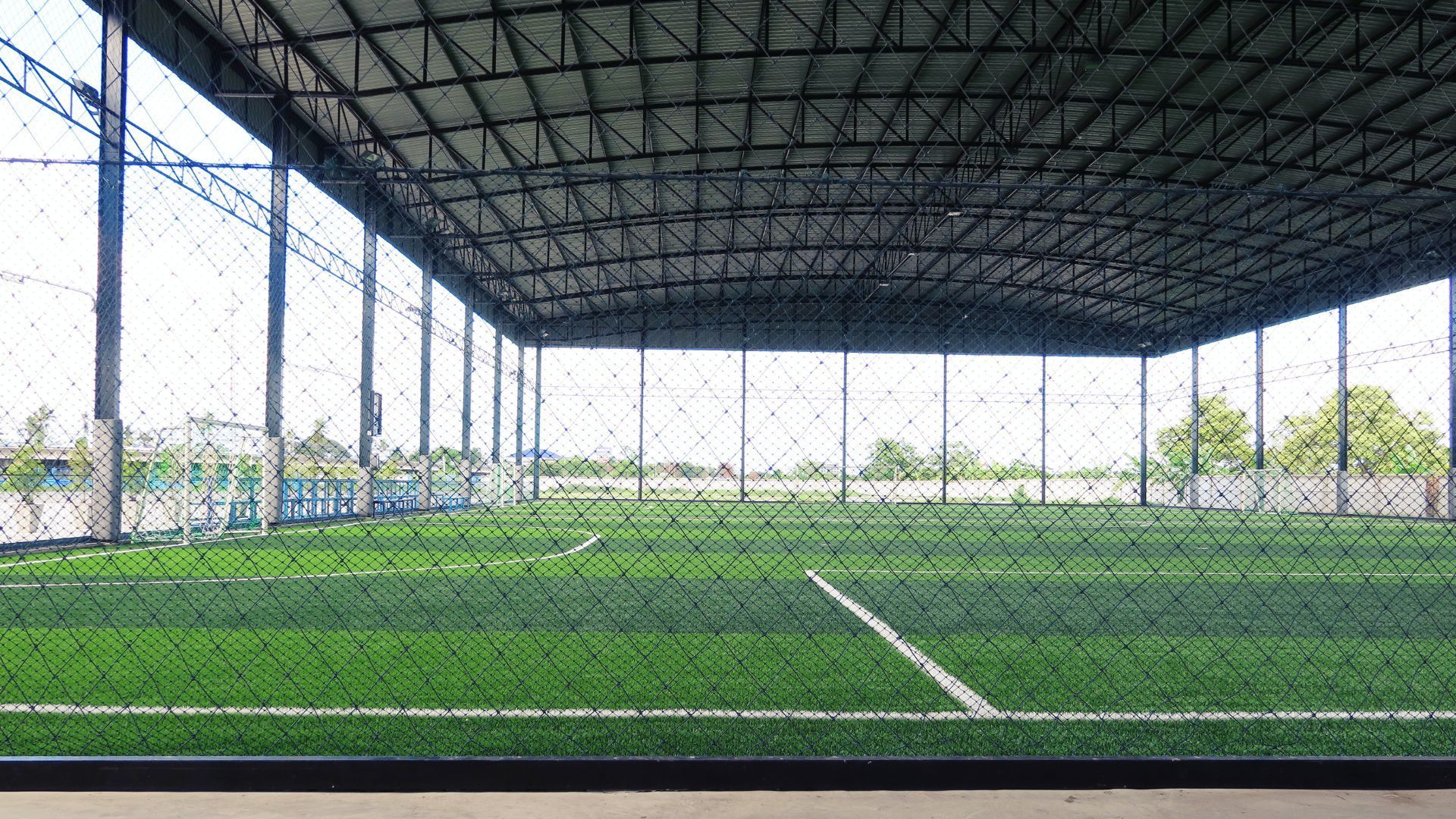 Terrain de football en salle avec gazon synthétique vert, lignes blanches et un haut toit métallique, visible à travers une clôture en grillage.