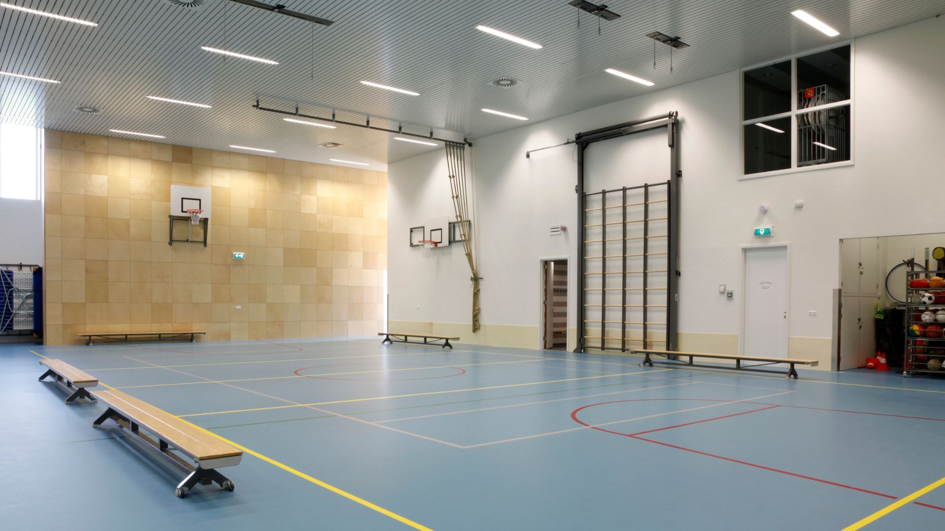 Une salle de sport intérieure avec un sol bleu clair, des murs en bois, des paniers de basket, une corde d'escalade et de longs bancs en bois.