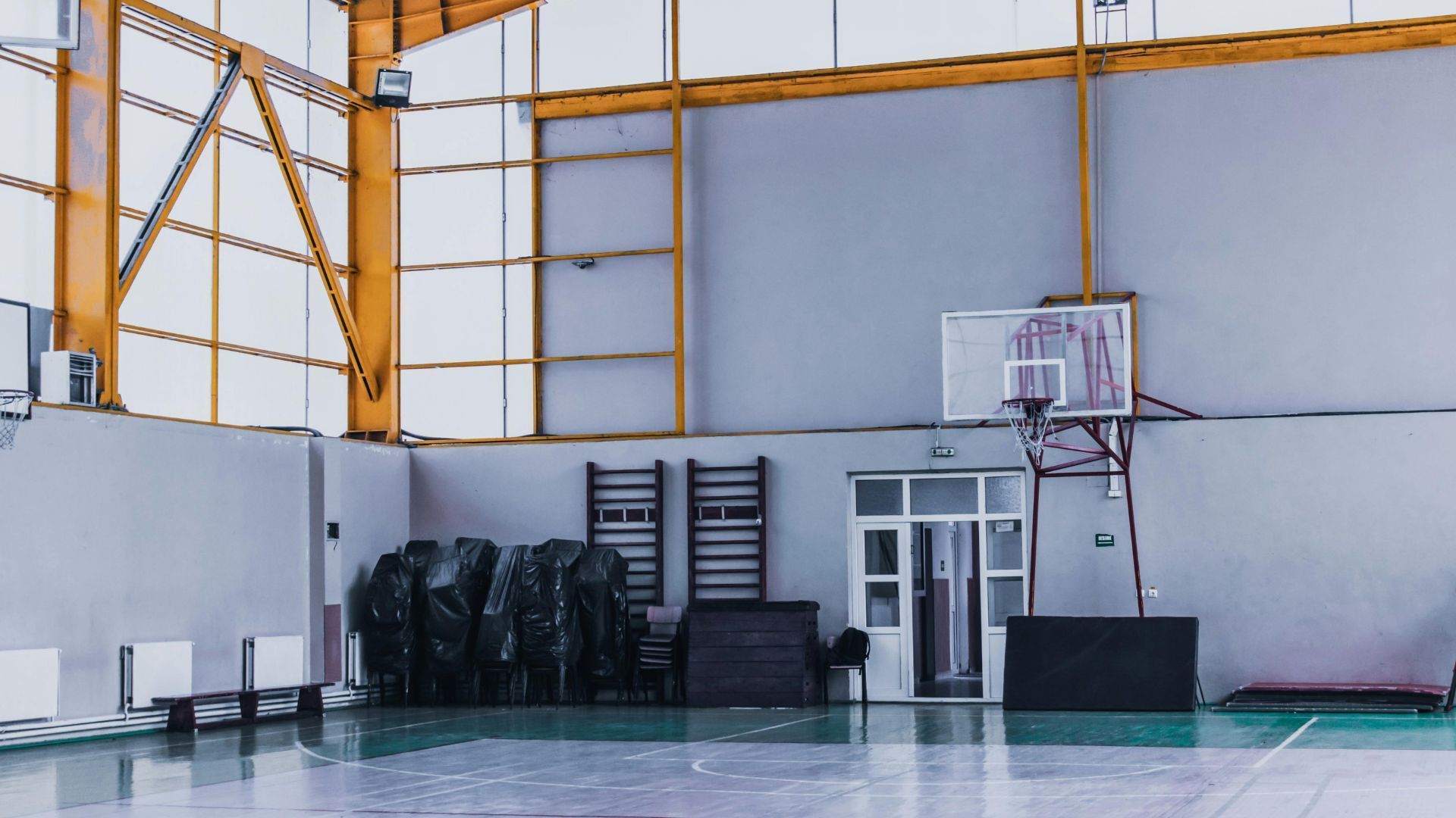 Une salle de sport intérieure avec un panier de basket sur un mur gris, des poutres métalliques jaunes et des chaises empilées sur le côté.