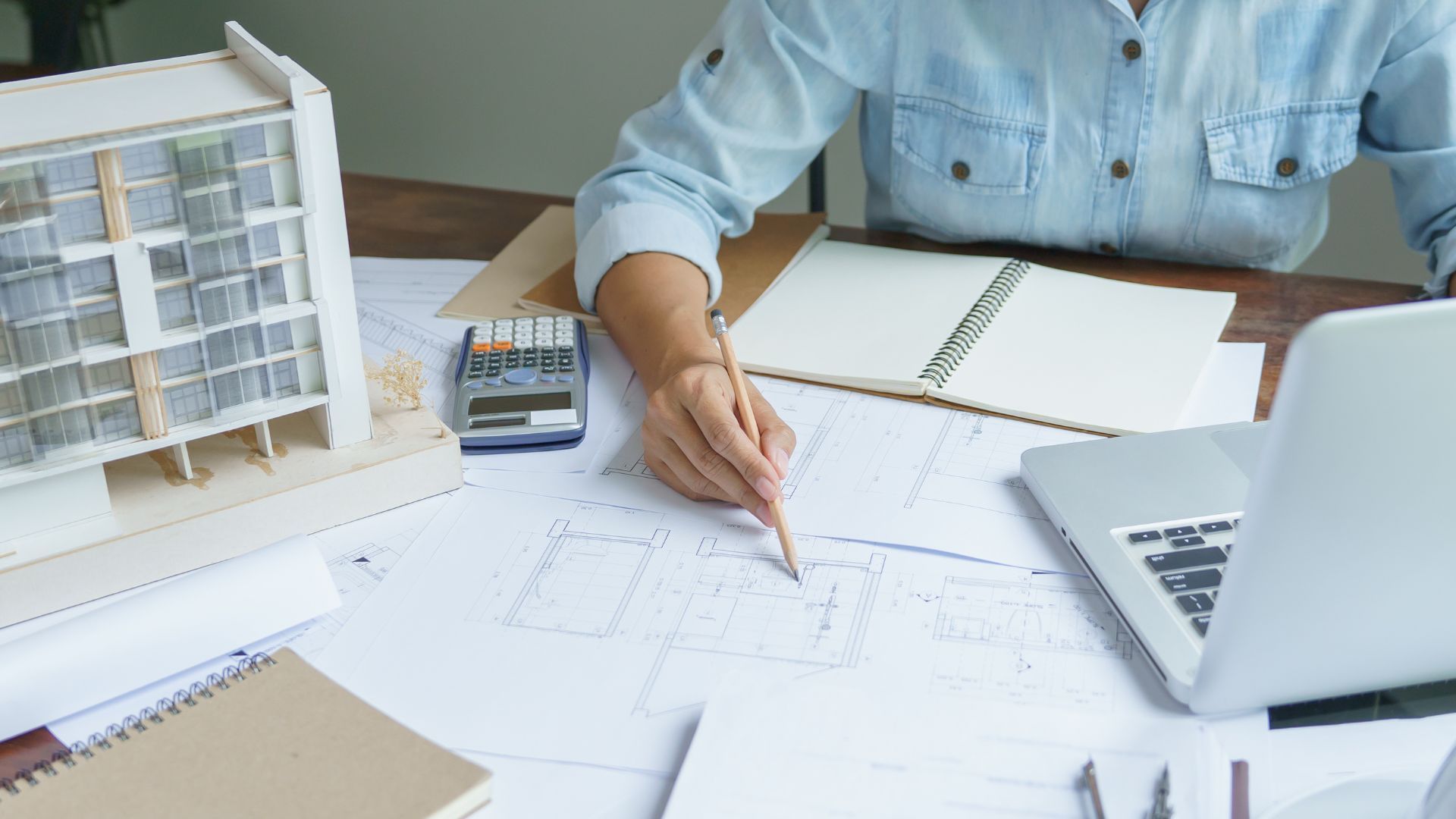 Un architecte travaillant à son bureau avec des plans de construction, une maquette, une calculatrice et un ordinateur portable.
