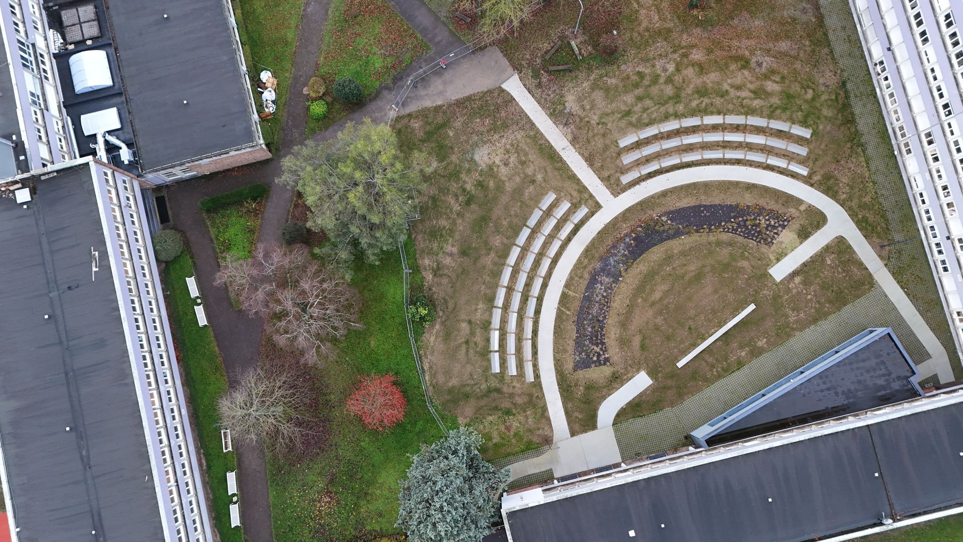 Une vue aérienne révèle un amphithéâtre extérieur avec des gradins semi-circulaires nichés entre des bâtiments et des espaces verts.