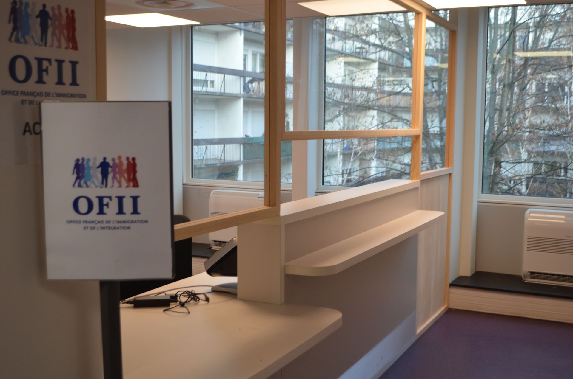 Un espace d'accueil de bureau avec un panneau OFII sur un support, comprenant un comptoir avec une cloison vitrée à cadre en bois.