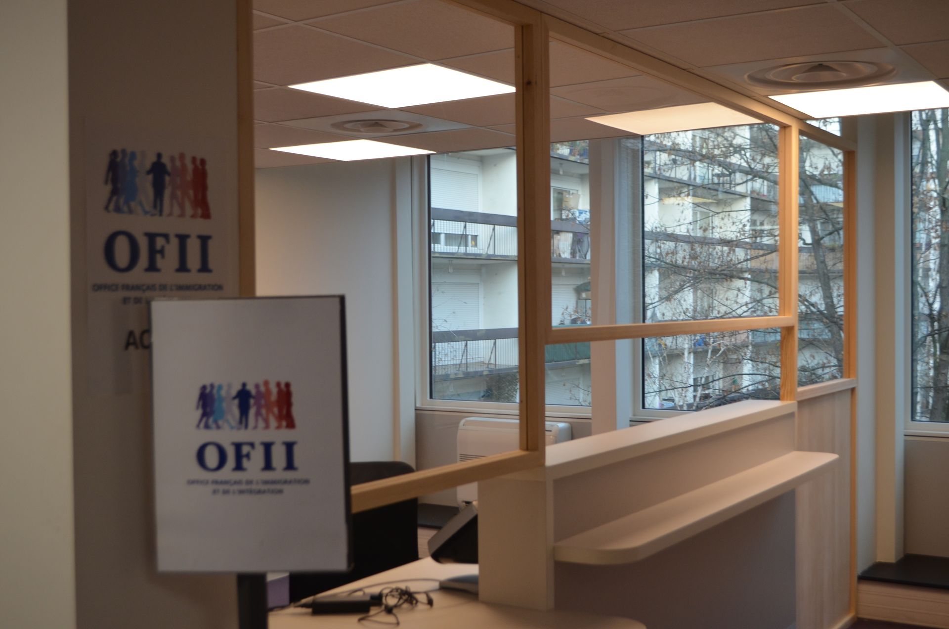 Un espace d'accueil avec un panneau portant le logo de l'OFII sur un support et une cloison vitrée séparant l'espace de travail de la vue extérieure.