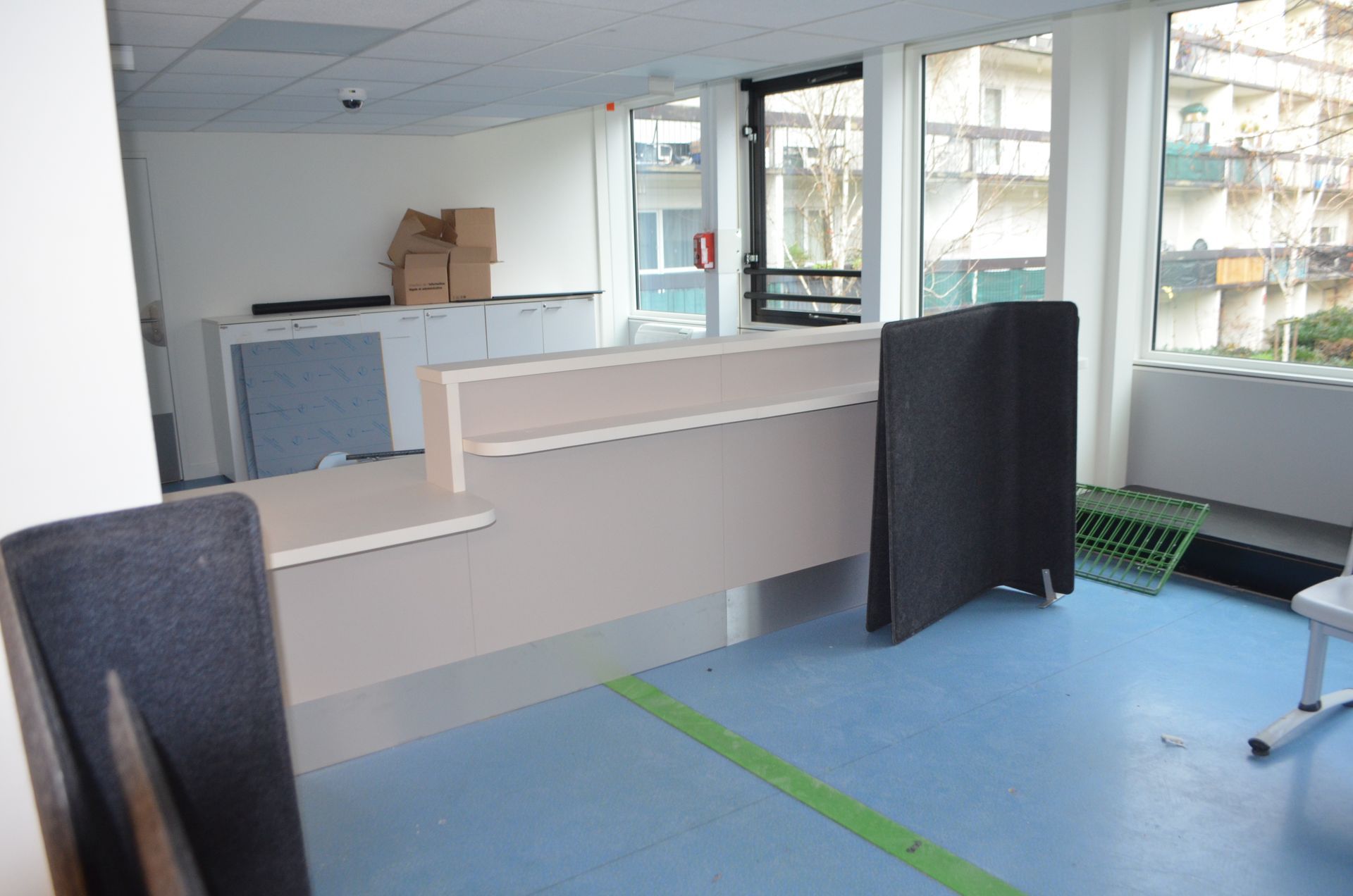 Un comptoir d'accueil clair, avec des cloisons grises, se trouve dans un espace de bureaux au sol bleu et aux grandes fenêtres.