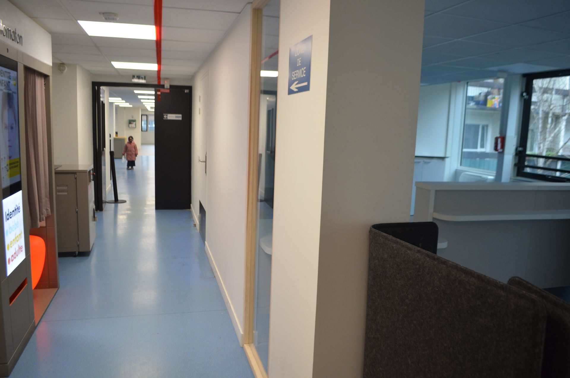 Un couloir d'hôpital avec un sol bleu, une borne libre-service sur la gauche et une personne qui marche au loin.