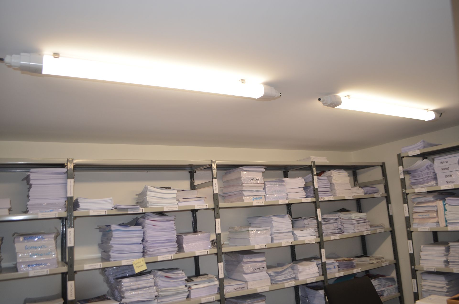 Des étagères métalliques remplies de piles de papiers, éclairées par deux lampes fluorescentes vives fixées au plafond blanc.
