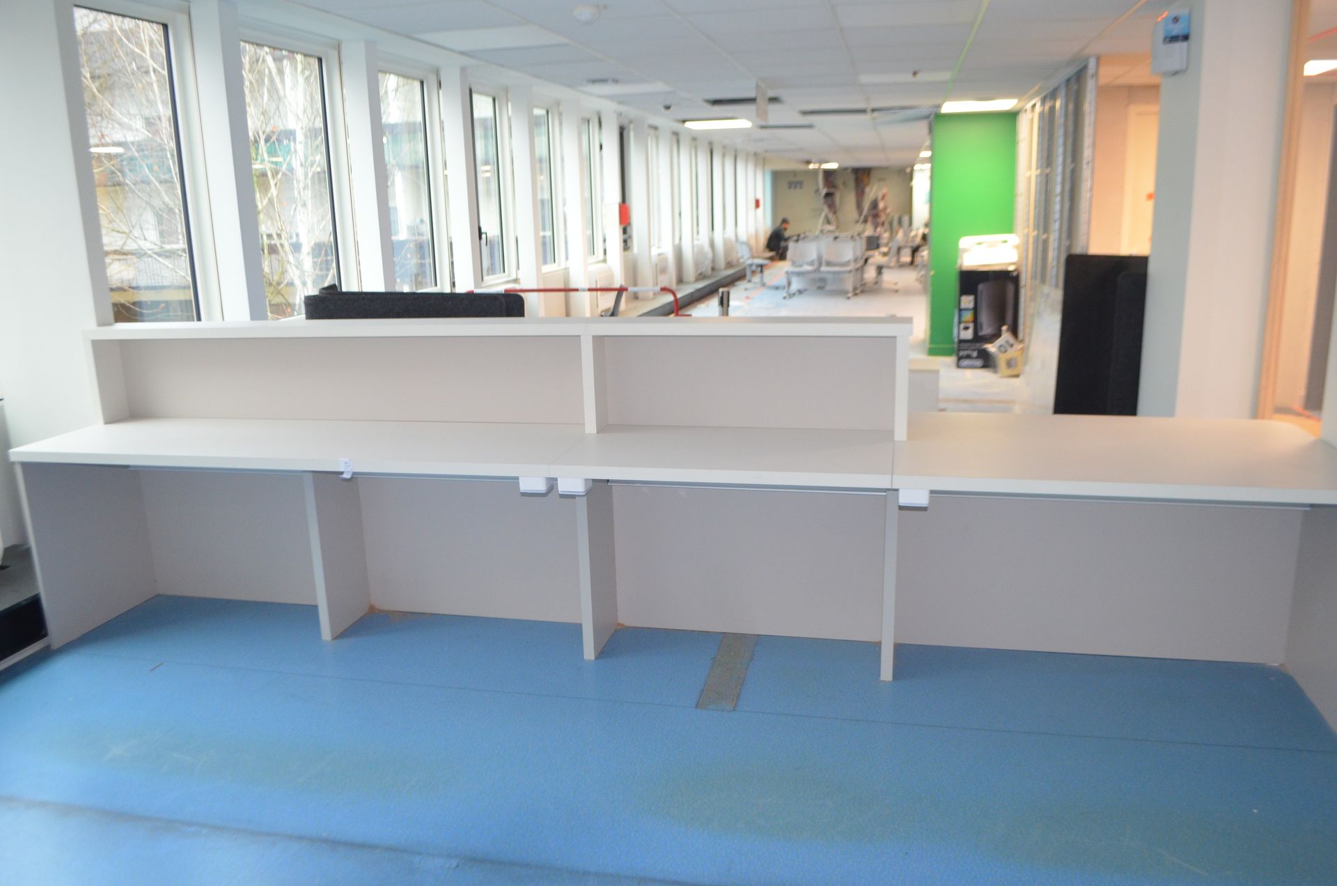 Un comptoir d'accueil clair est installé sur un sol bleu dans un espace de bureaux doté de grandes fenêtres et d'un mur vert au fond.