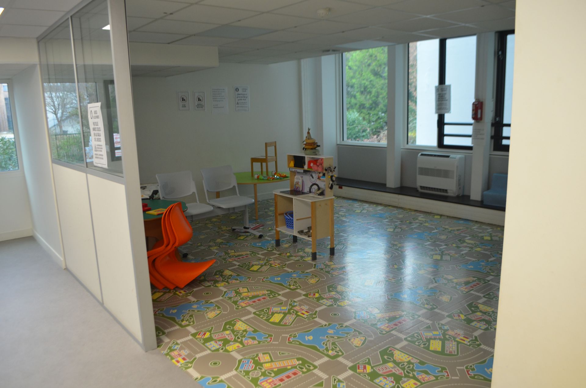Un espace de jeux pour enfants avec un tapis représentant une carte de la ville, des petites chaises orange et blanches et une étagère en bois avec des jouets.
