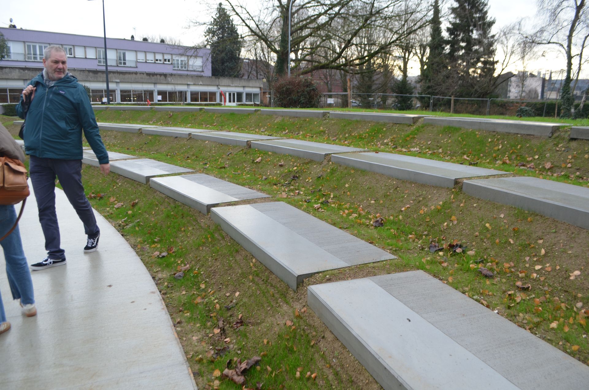 Une personne marche le long d'un chemin en béton qui longe une esplanade herbeuse en gradins avec des marches en béton, dans un parc.