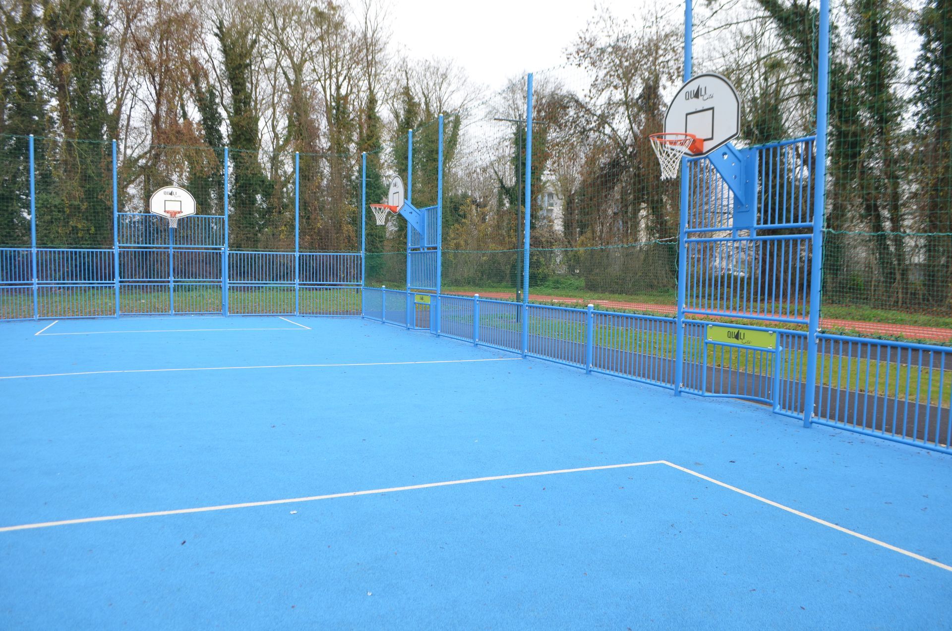 Terrain de sport extérieur bleu avec plusieurs paniers de basket et une clôture, entouré d'arbres par temps couvert.