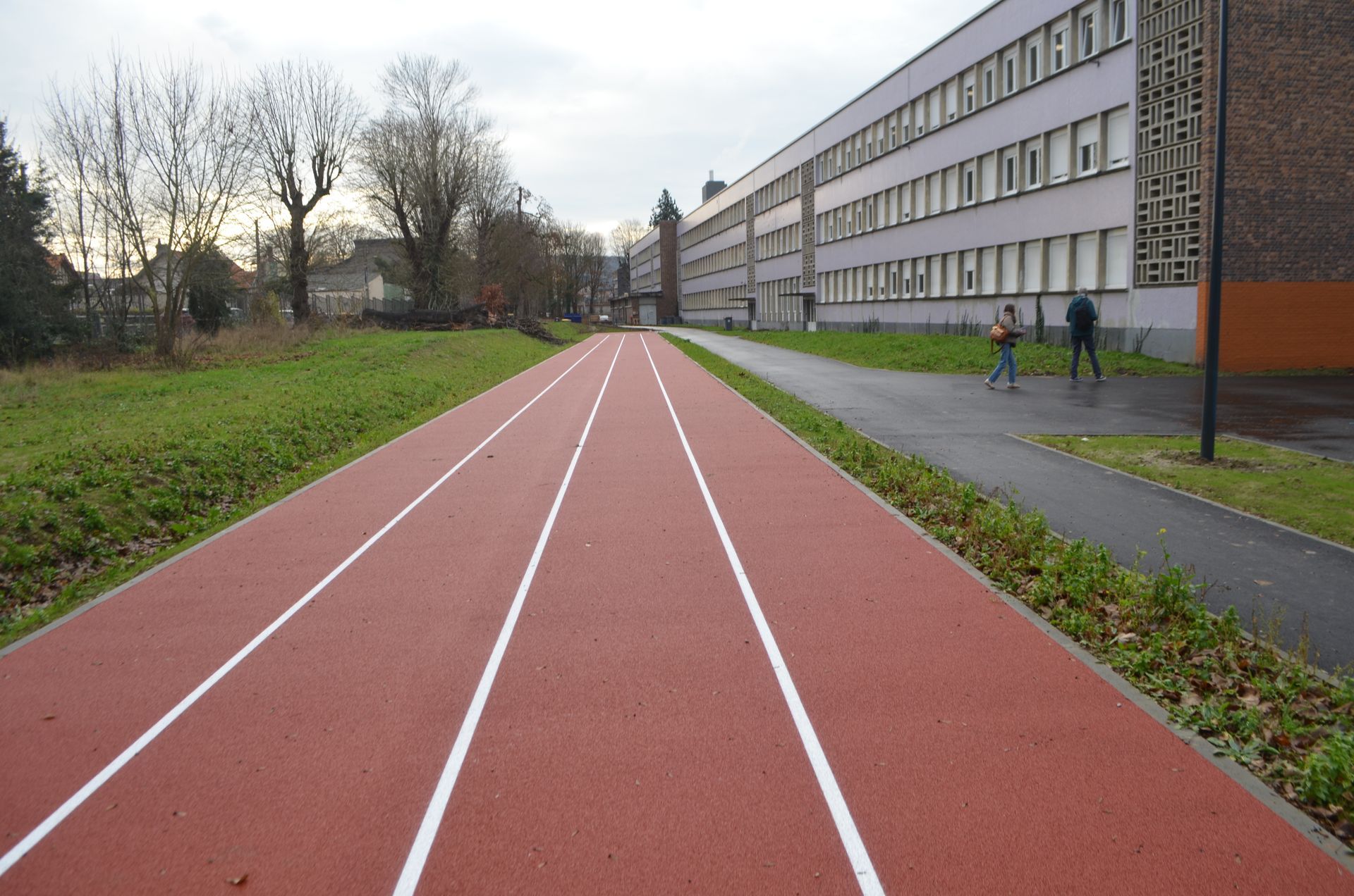 Une piste d'athlétisme rouge, délimitée par des lignes blanches, longe un long bâtiment scolaire à plusieurs étages doté d'une allée pavée.