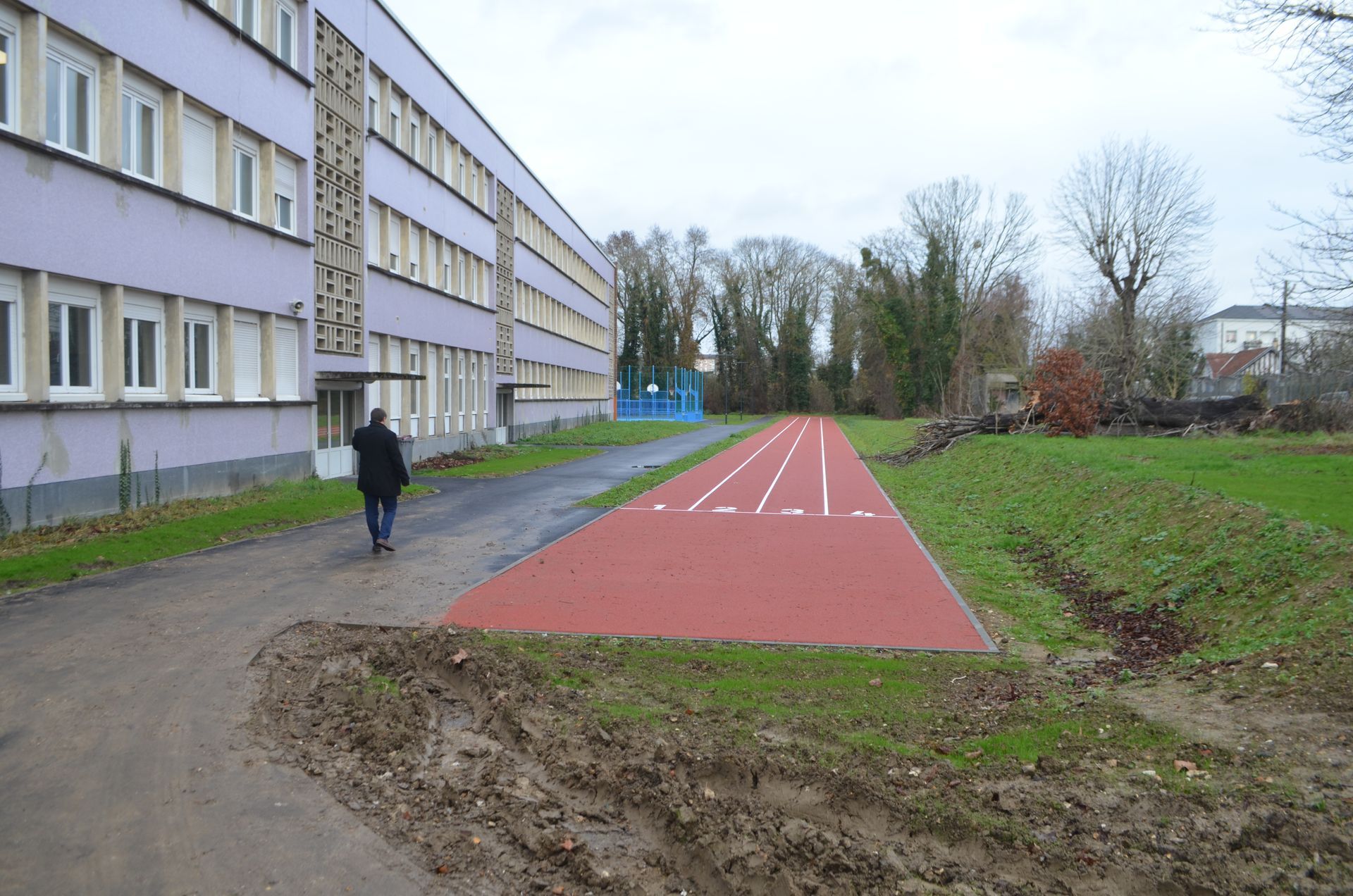 Une personne marche sur un chemin longeant un bâtiment scolaire moderne et une nouvelle piste d'athlétisme rouge, en plein air.