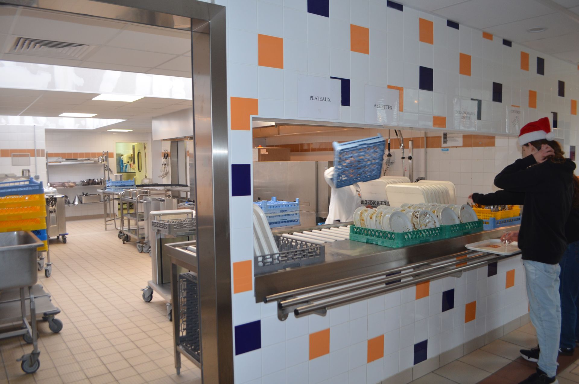 Une personne portant un bonnet de Père Noël se tient au comptoir d'une cafétéria, avec des plateaux et du matériel de restauration en arrière-plan.