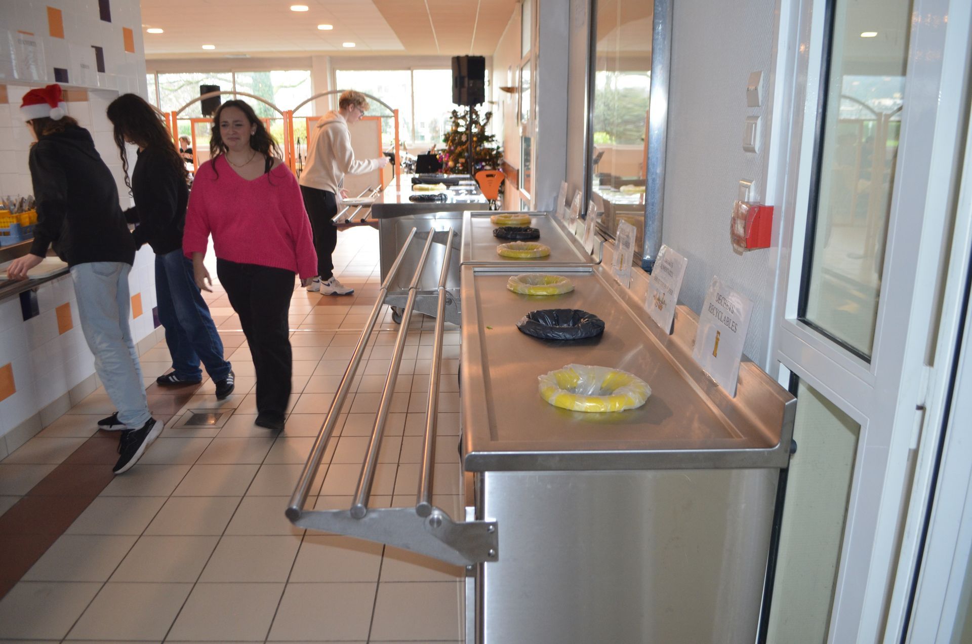 Dans une cafétéria moderne et lumineuse, des personnes se tiennent près d'un comptoir de service en acier inoxydable où sont présentés divers plats.