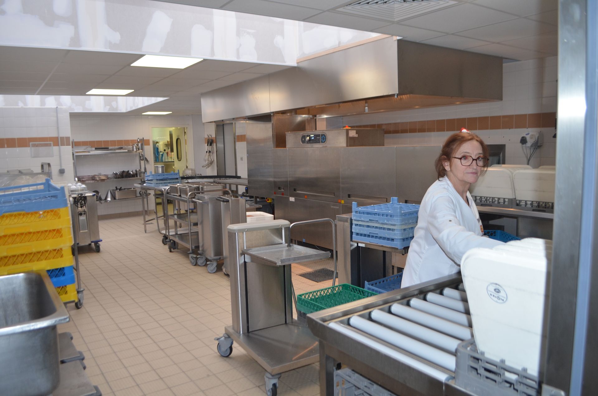 Une personne portant une blouse blanche travaille dans une cuisine professionnelle équipée de matériel en acier inoxydable, de chariots et de caisses en plastique.