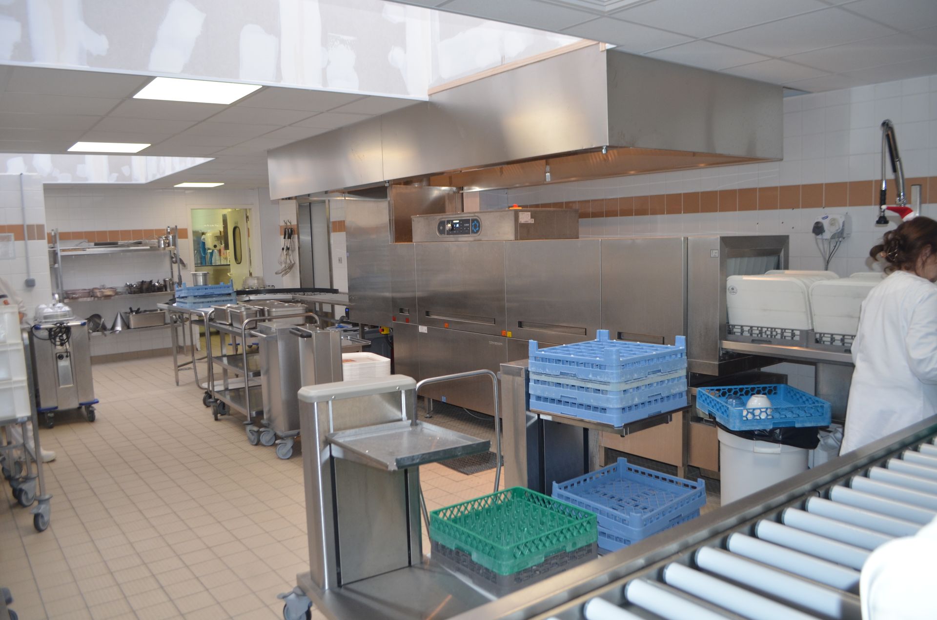 Poste de lavage de vaisselle industriel dans une cuisine commerciale, avec des caisses en plastique bleues et vertes sur des étagères et des convoyeurs métalliques.