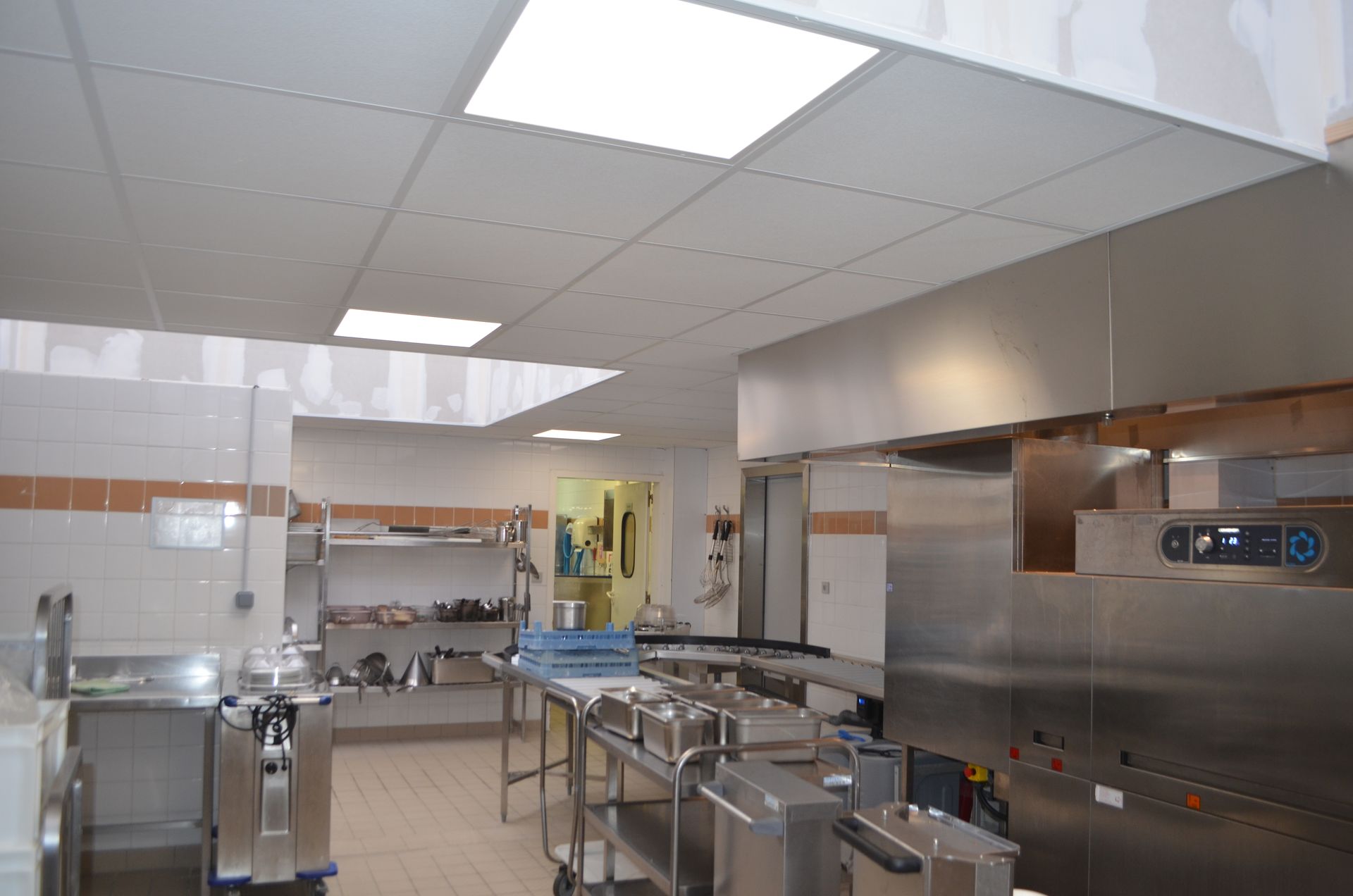 Une cuisine professionnelle en acier inoxydable avec étagères, tables de préparation et éclairage encastré lumineux au plafond.