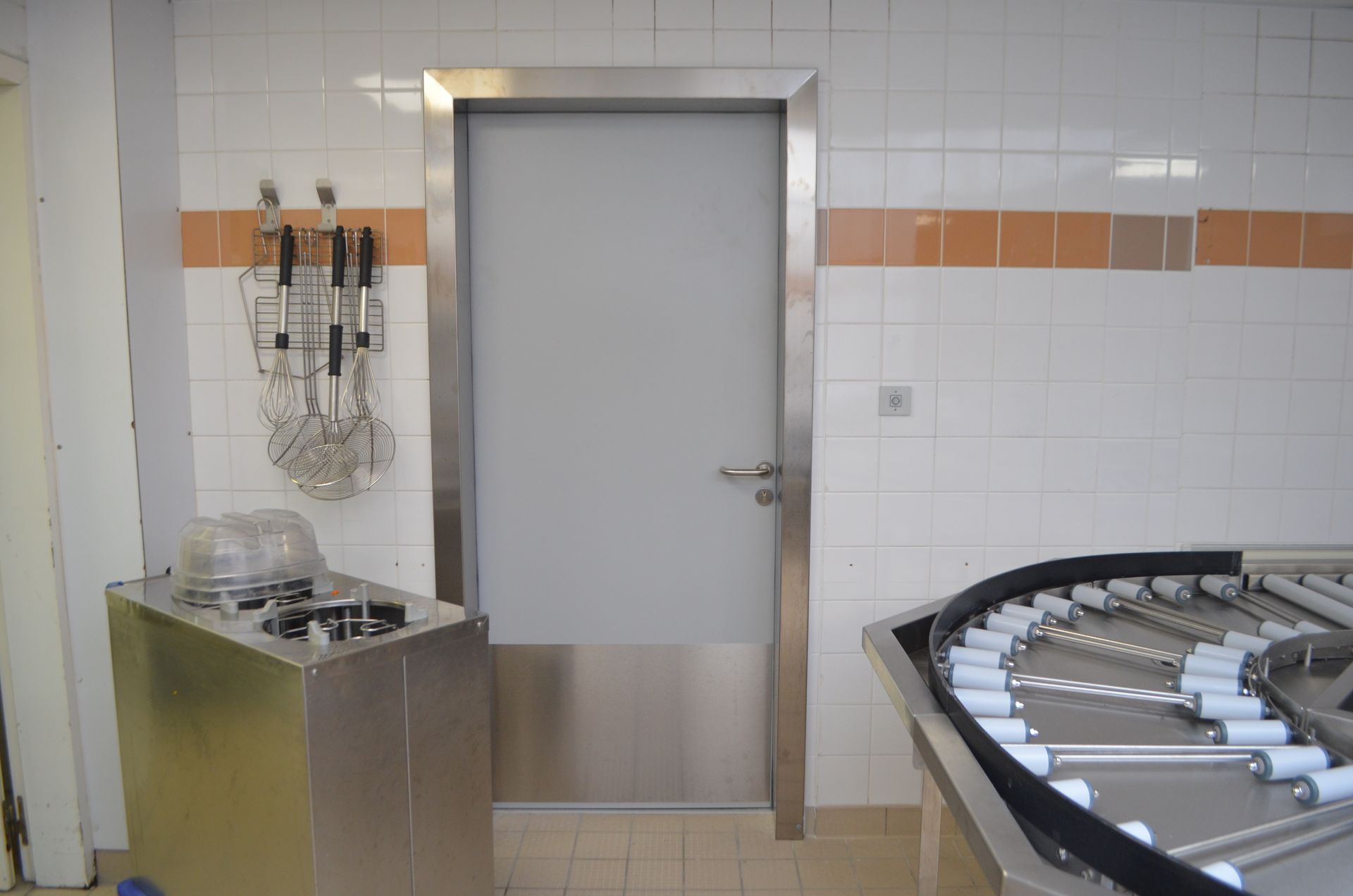 Une porte grise à cadre métallique dans une cuisine industrielle carrelée, à côté d'ustensiles suspendus et d'un système de convoyeur.