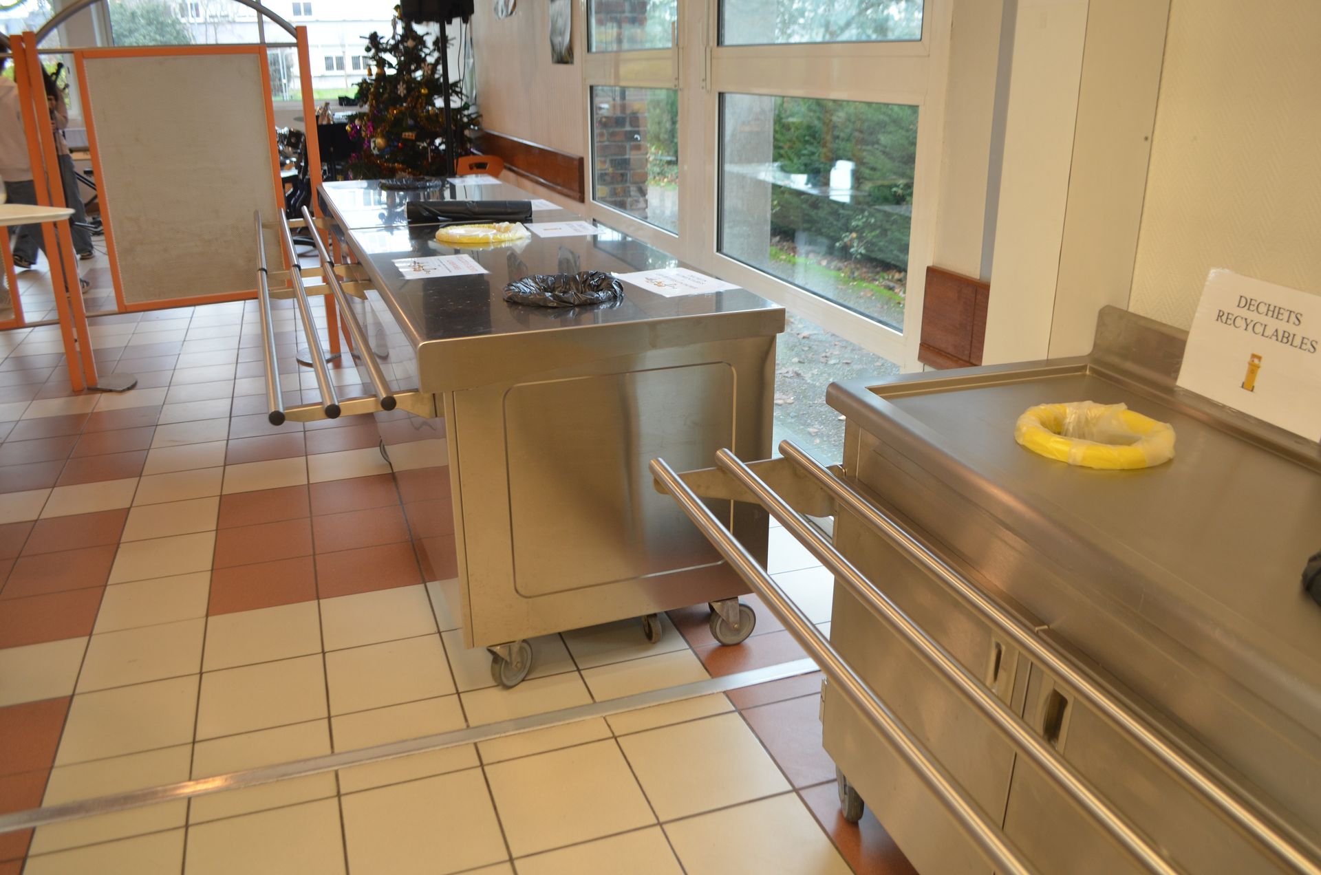Des tables de buffet en acier inoxydable sont disposées dans une pièce lumineuse au sol carrelé, offrant une vue sur un espace extérieur.