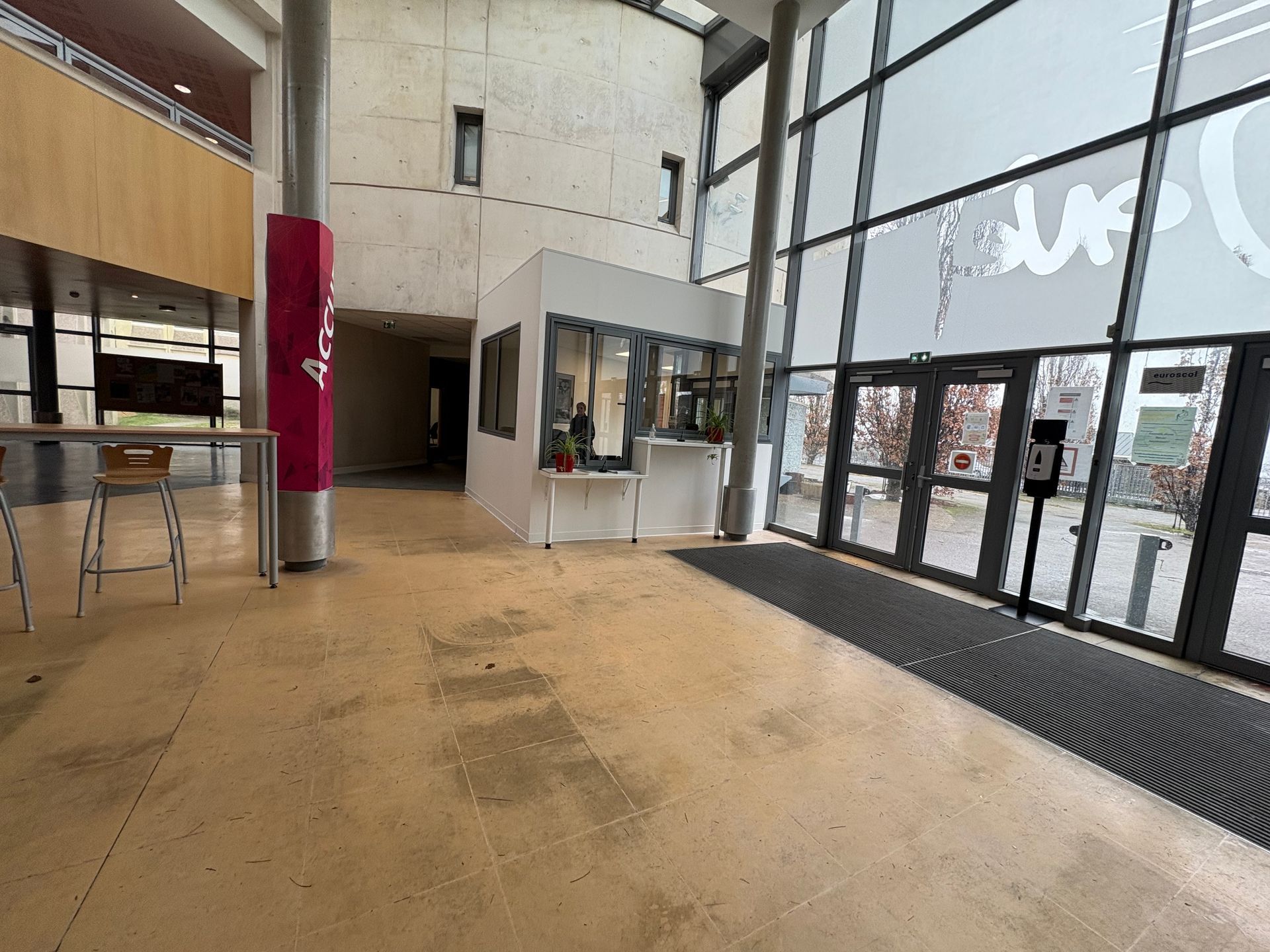Vue panoramique du hall d'un immeuble lumineux, avec un sol poli, une guérite, des portes vitrées et un haut pilier.