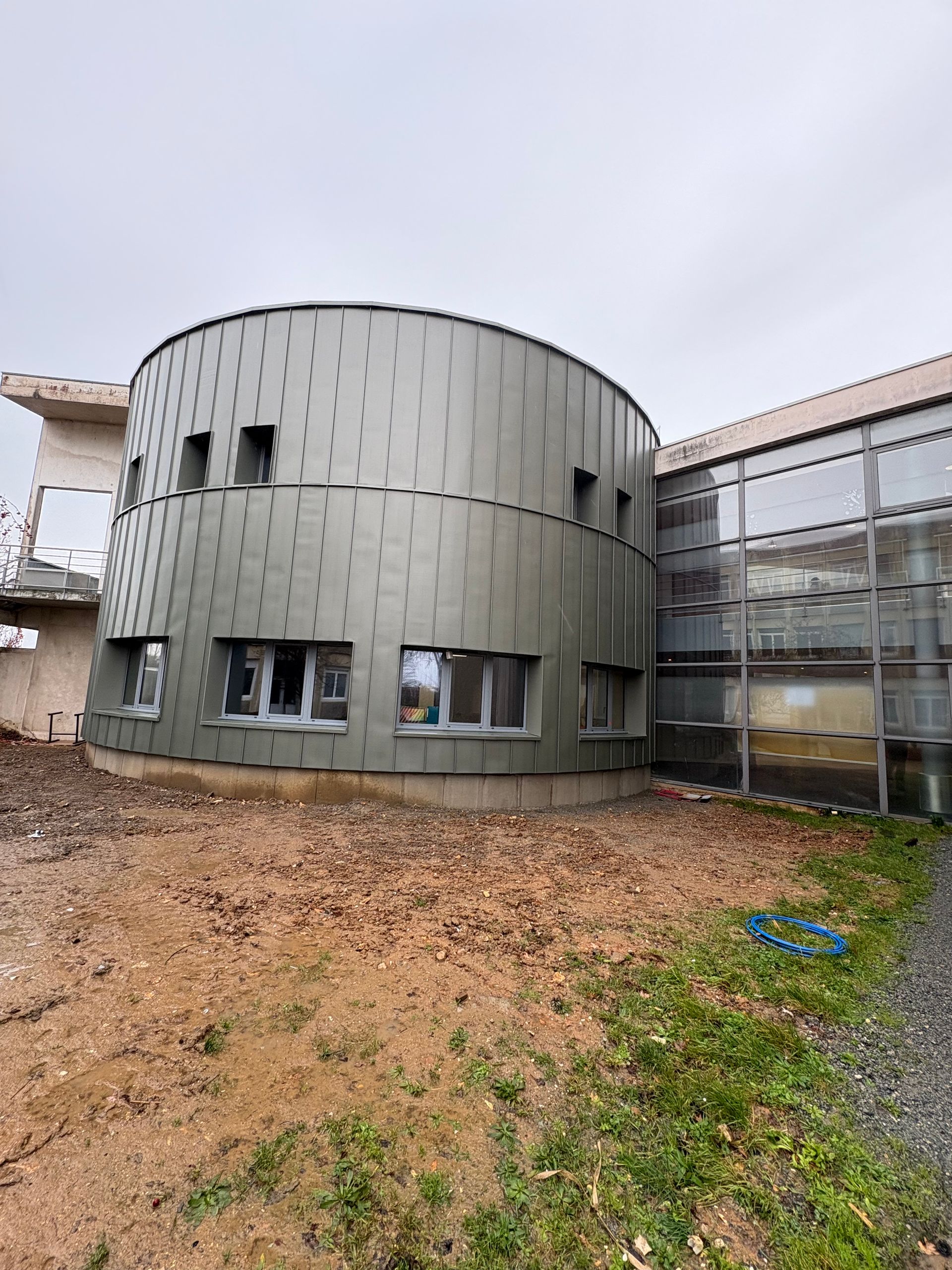 Un bâtiment cylindrique aux panneaux métalliques et percé de plusieurs fenêtres se dresse à côté d'une structure aux parois de verre, sous un ciel gris.