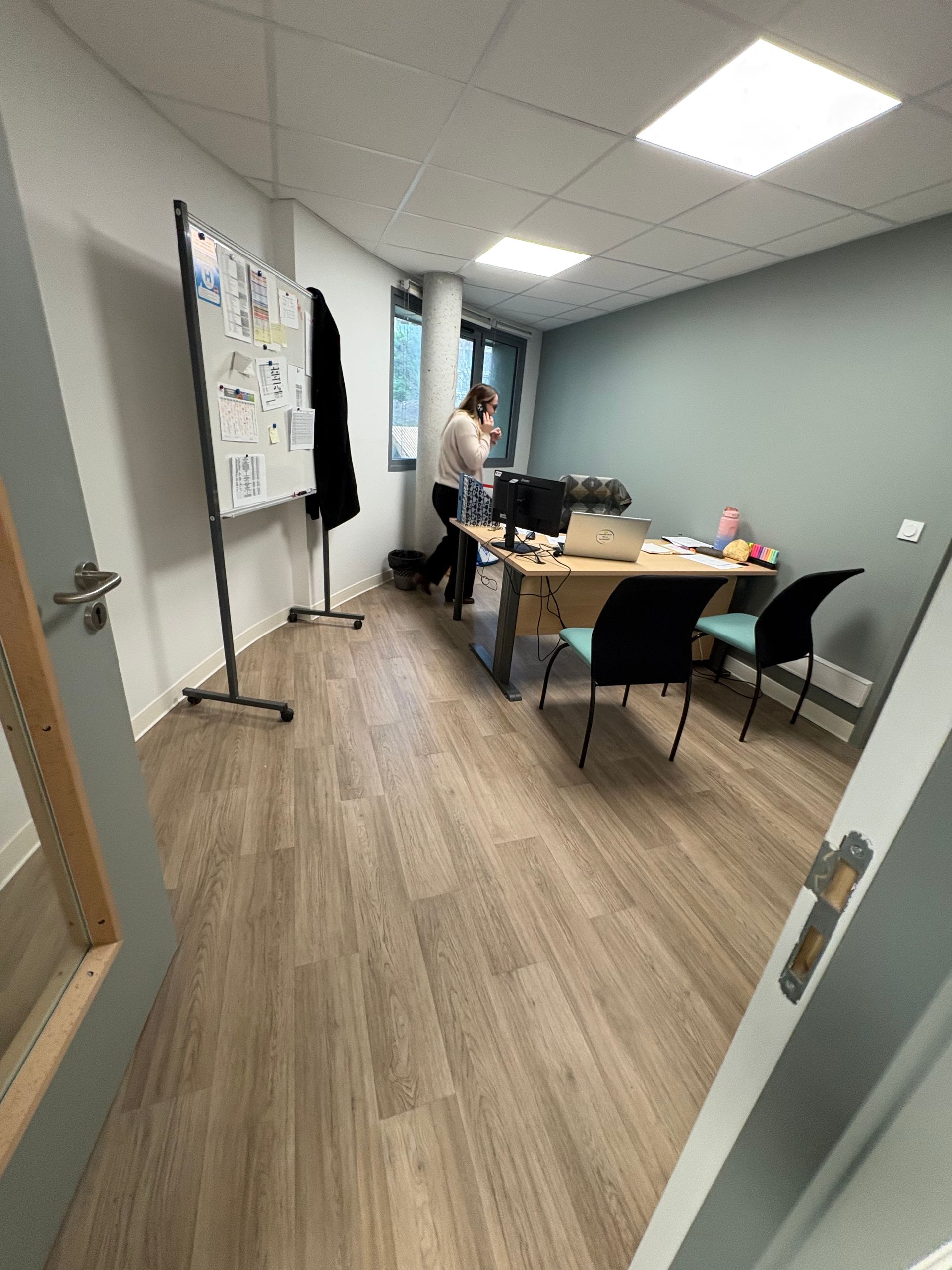 Un bureau avec un sol imitation bois, un tableau blanc roulant et un bureau avec des chaises, avec une personne debout près d'une fenêtre.