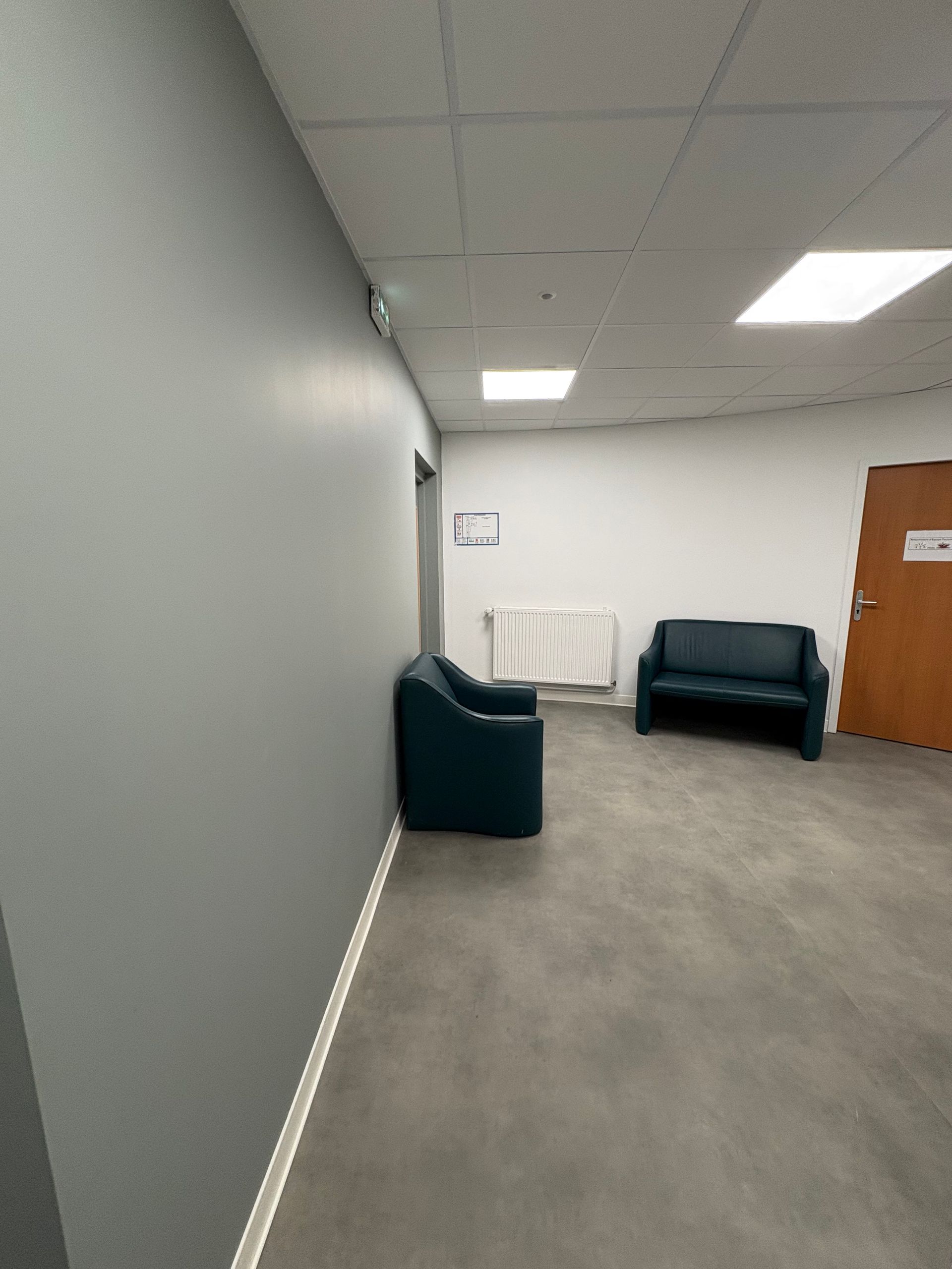 Une salle d'attente avec deux chaises bleu foncé contre un mur gris, un radiateur blanc et une porte en bois.