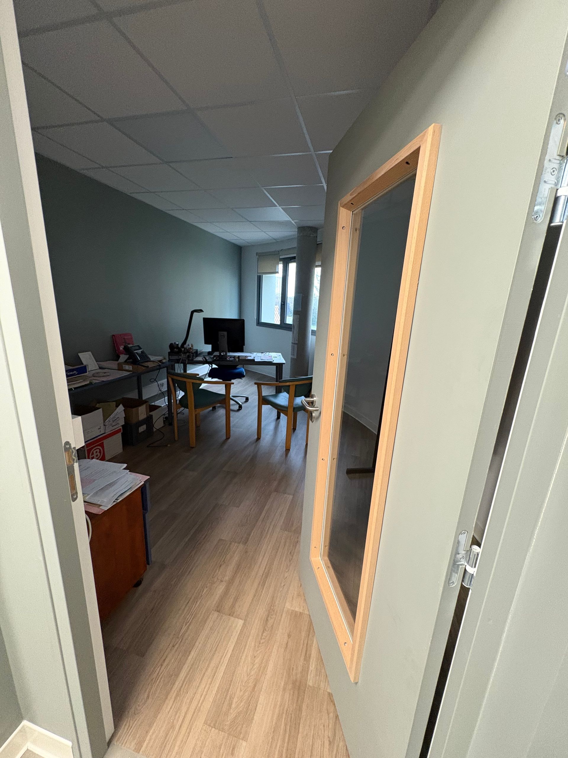 Vue à travers une porte entrouverte donnant sur un bureau avec parquet, bureau, chaises et grandes fenêtres.