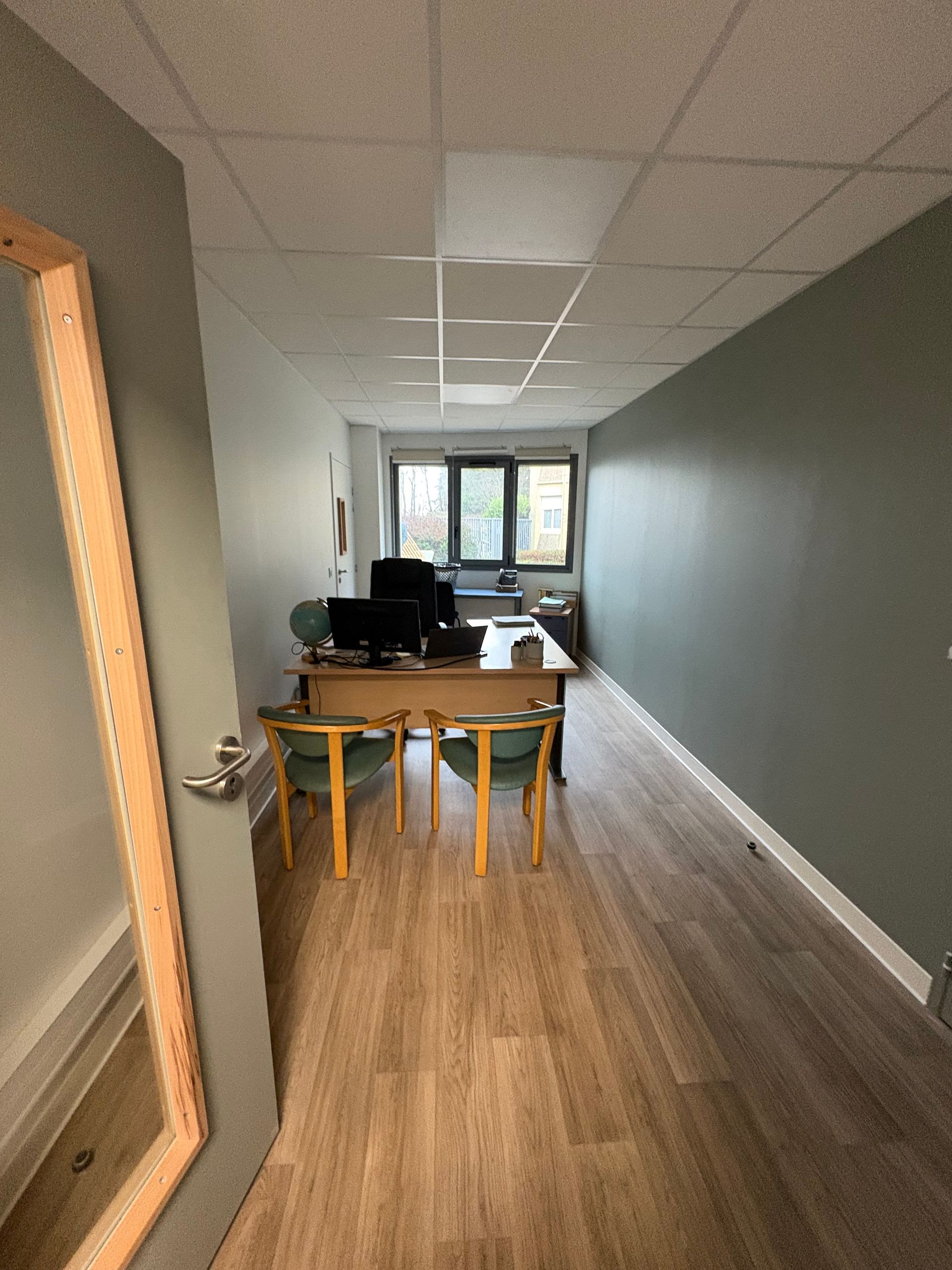 Un espace de bureau étroit et vide, avec un parquet clair, un bureau en bois avec un ordinateur, deux chaises et une fenêtre au fond.