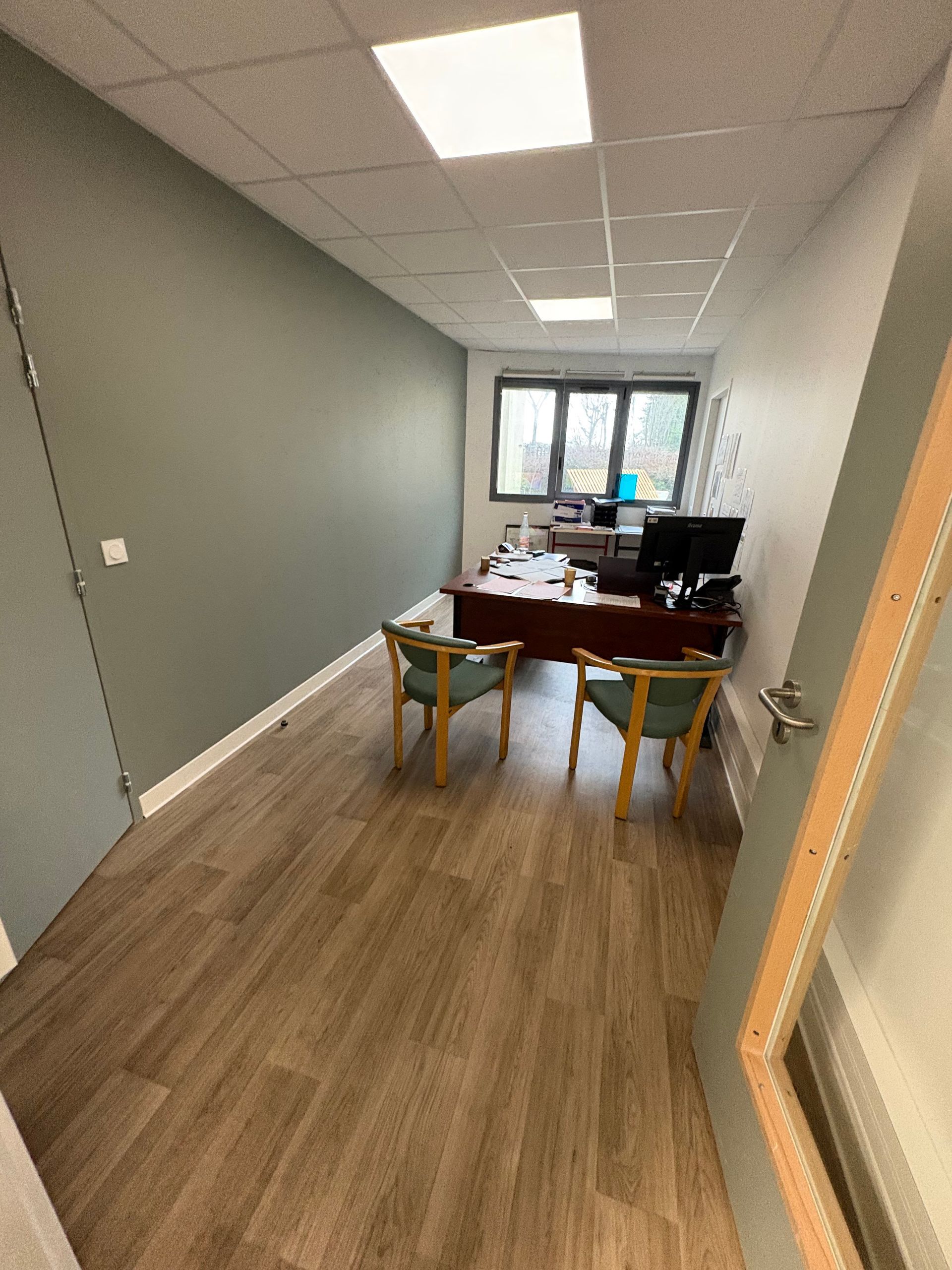 Petit bureau aux murs gris, sol imitation bois, un bureau avec un ordinateur et deux chaises face à celui-ci, près d'une fenêtre.