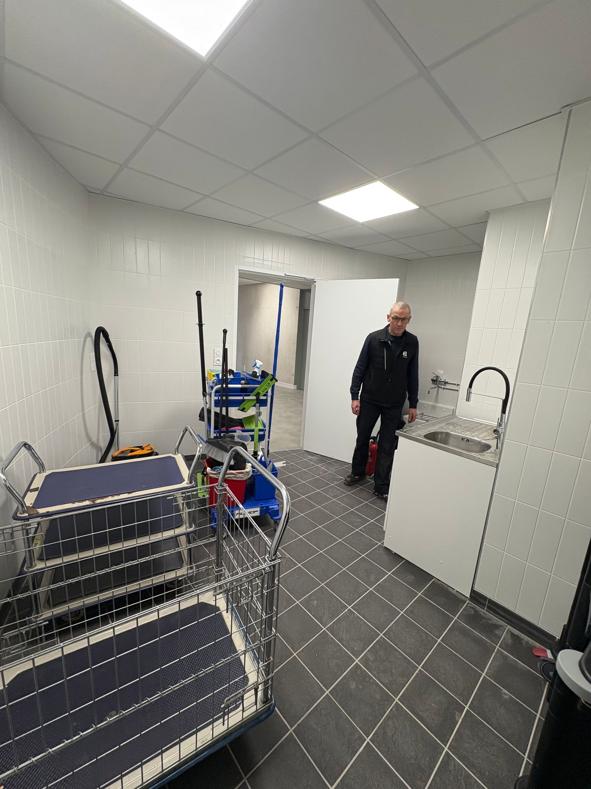 Une personne se tient dans un local technique avec des chariots de nettoyage, un évier et un sol carrelé foncé.