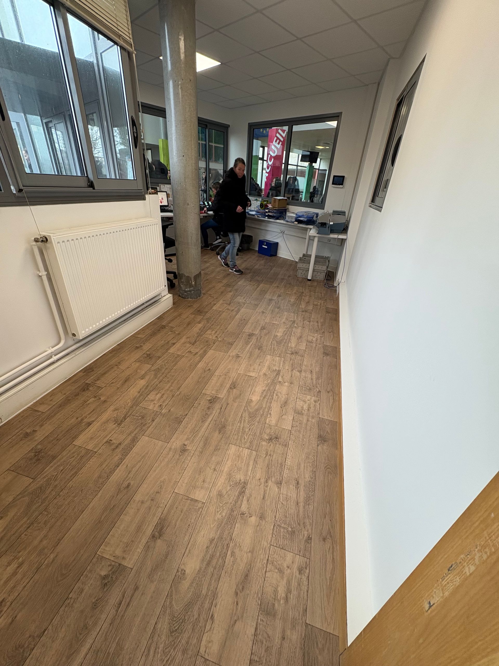 Une personne vêtue de sombre traverse un espace de bureau avec un sol en parquet, des murs blancs et une grande fenêtre.