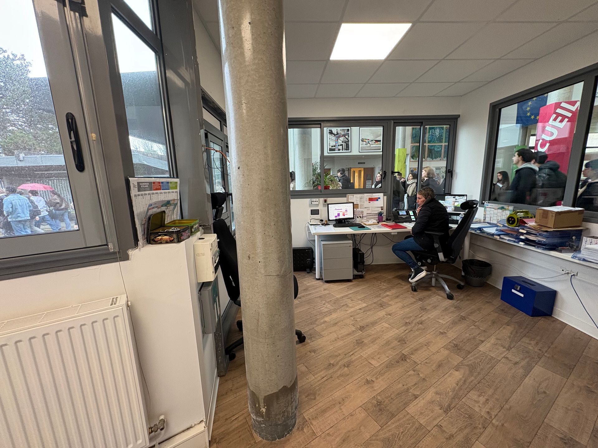 Une personne est assise à un bureau dans un bureau lumineux avec un sol imitation bois, un grand pilier et des fenêtres donnant sur l'extérieur.