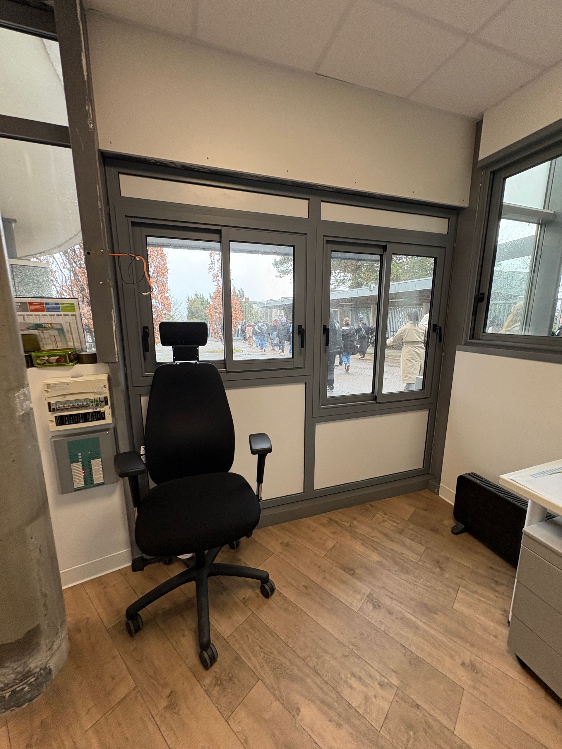 Une chaise de bureau noire dans une pièce moderne et lumineuse, avec des fenêtres donnant sur une allée et un parquet clair.