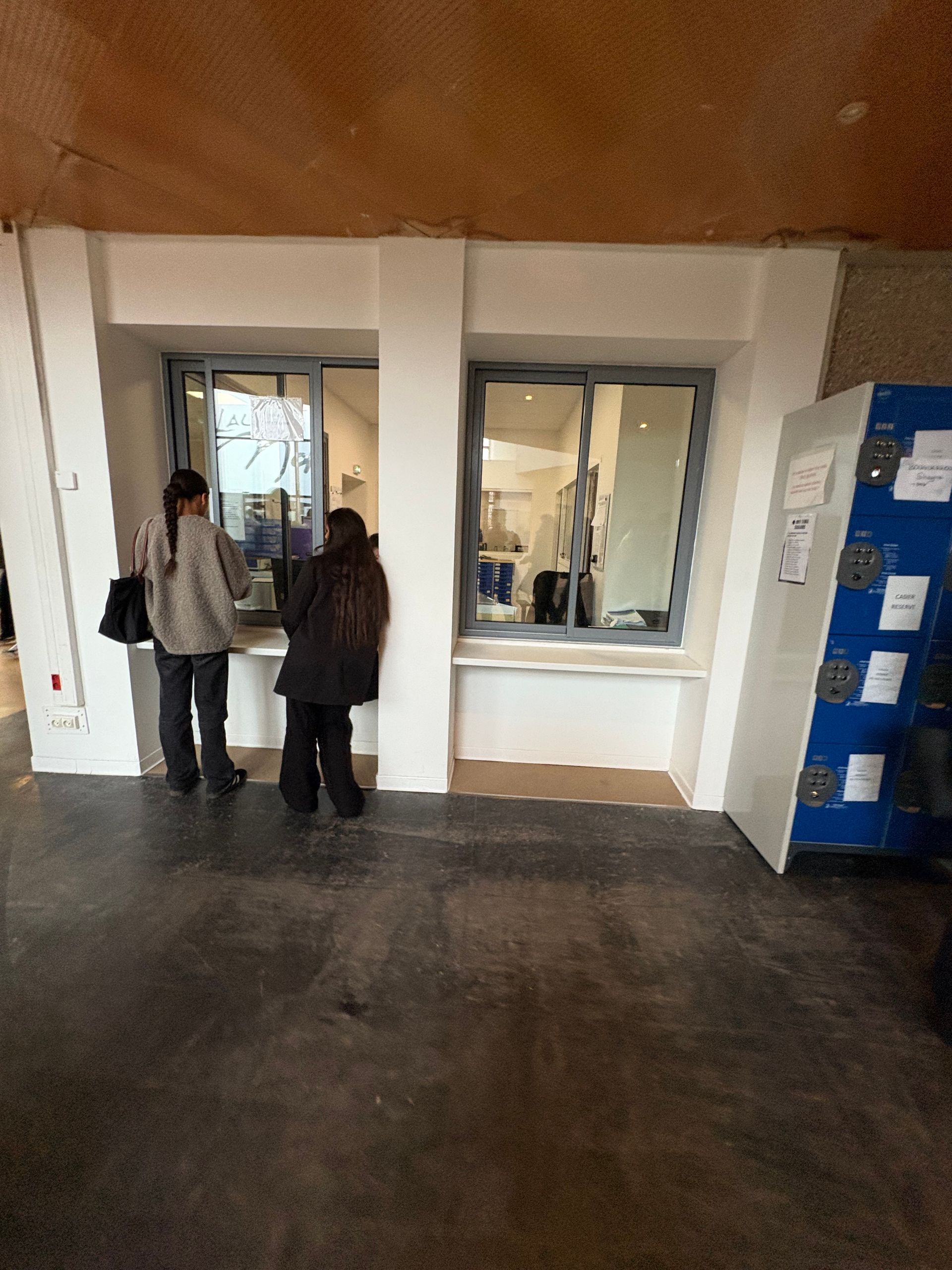 Deux personnes se tiennent devant un guichet de service dans un intérieur aux murs blancs et au sol en béton, à côté d'un casier de rangement bleu.
