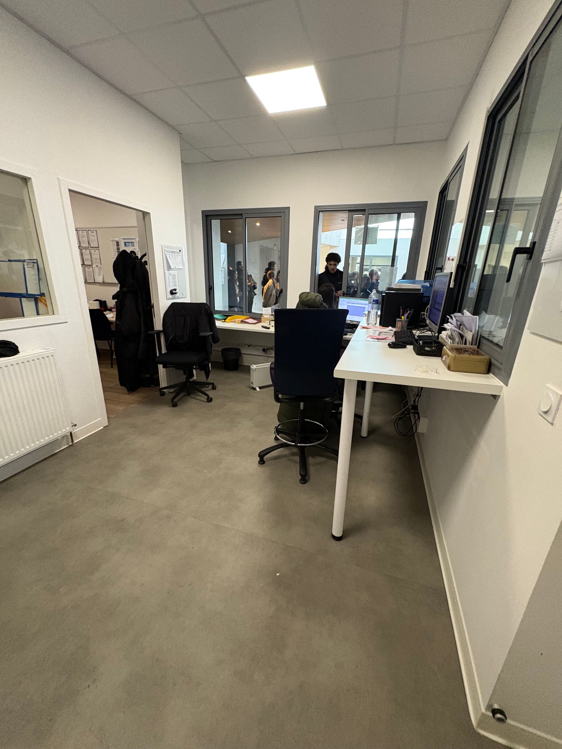 Un intérieur de bureau comprenant des bureaux, des chaises à roulettes et de grandes fenêtres donnant sur une pièce adjacente.