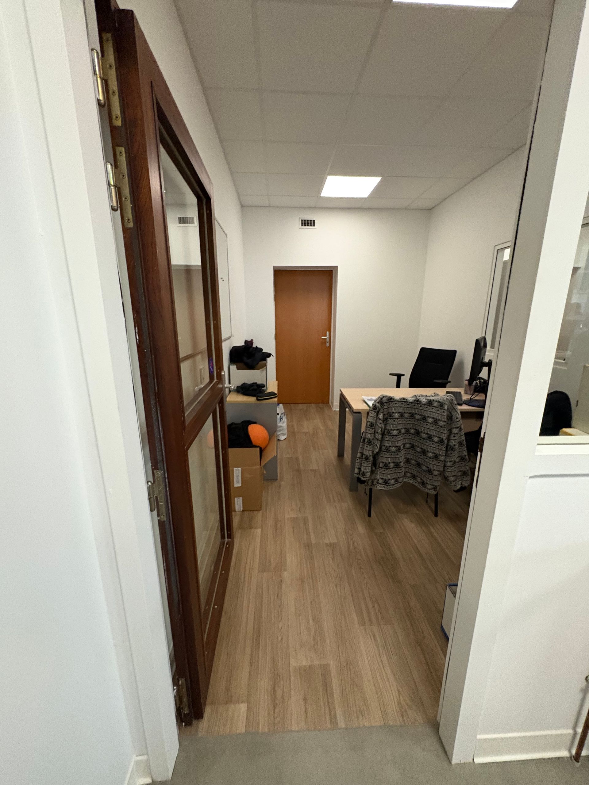 Une porte s'ouvre sur un bureau au parquet clair, avec un bureau et une chaise à motifs, et une porte fermée au fond.