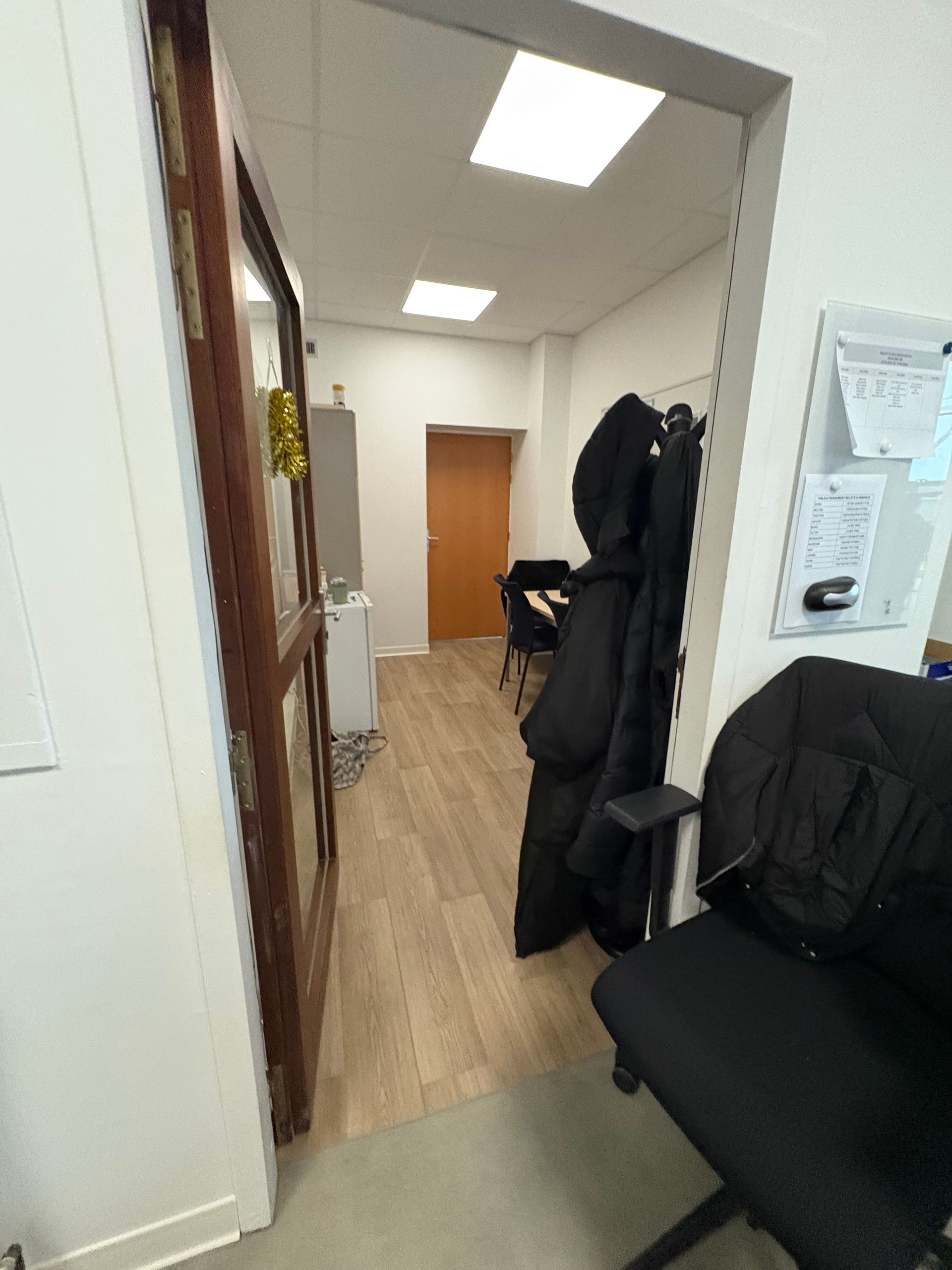 Vue à travers une porte ouverte sur un bureau lumineux avec un sol imitation bois, un bureau, un porte-manteau et une porte fermée au-delà.