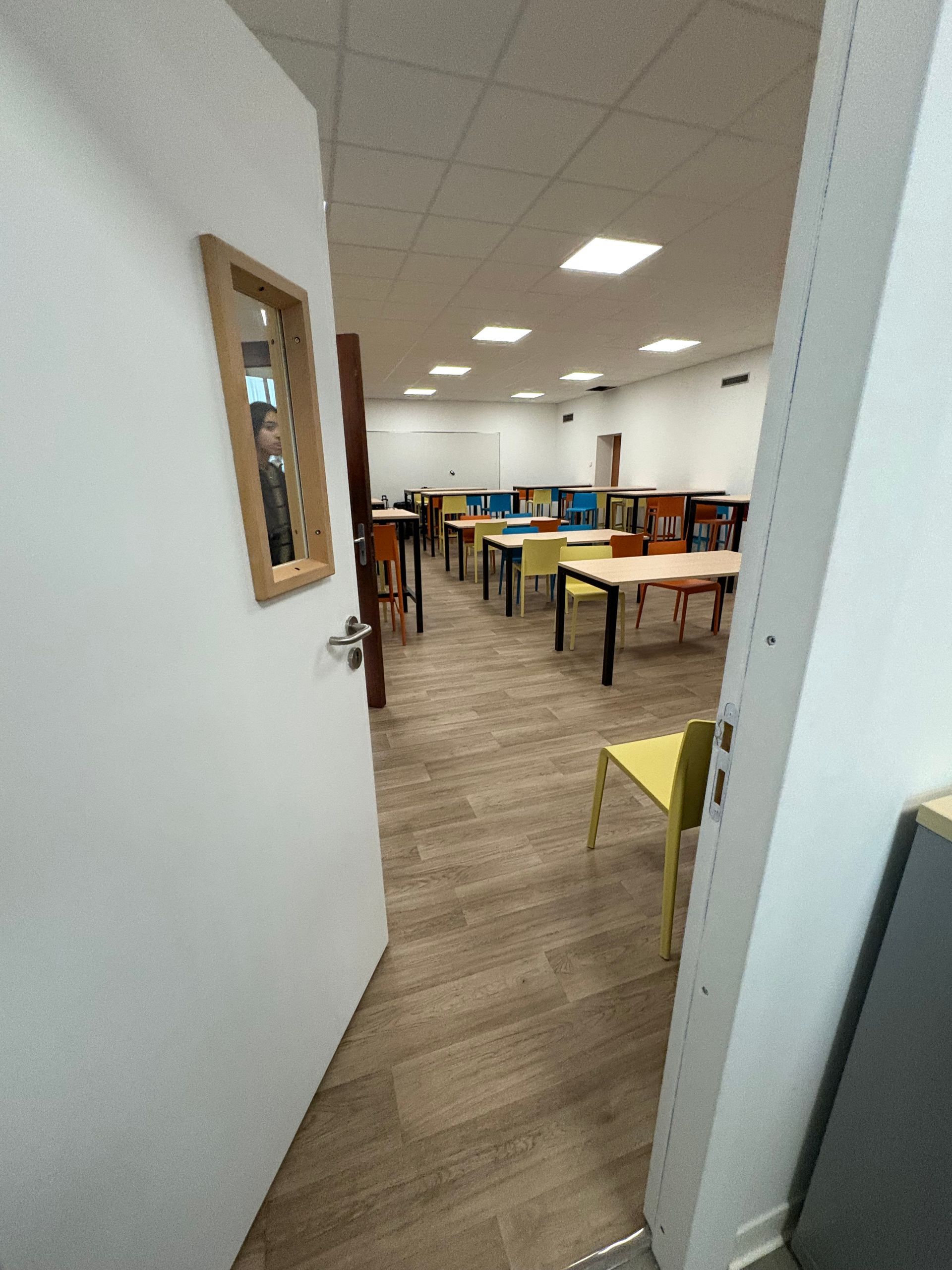 Vue, à travers une porte ouverte, d'une salle de classe lumineuse avec un sol en parquet, plusieurs tables et des chaises colorées.